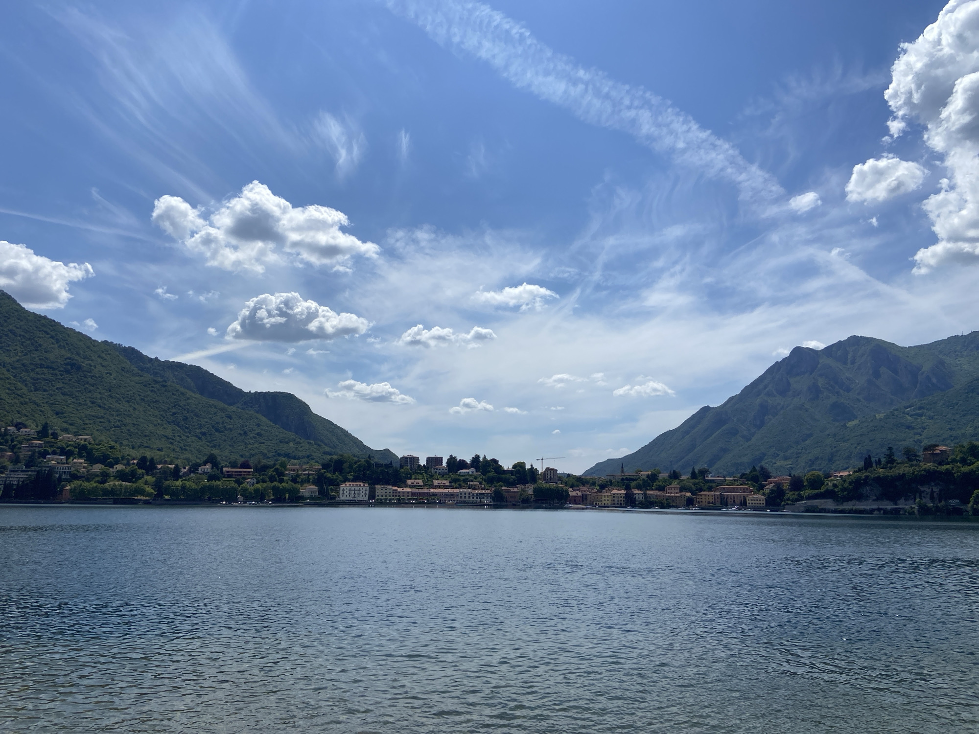 The image depicts a serene lake scene near the city of Lecco, Italy. The foreground is dominated by a large expanse of water with subtle ripples on its surface. In the midground, a line of buildings sits along the shoreline, possibly a town or village on the lake's edge. The buildings appear to be a mix of residential and possibly commercial structures with red-tiled roofs. The background features two prominent, densely forested mountains that frame the lake, adding depth and dimension to the scene. The sky is blue with a mix of puffy cumulus clouds and faint contrails, suggesting a clear day with air traffic. There are no people visible in the image. The overall environment suggests a calm and picturesque setting.