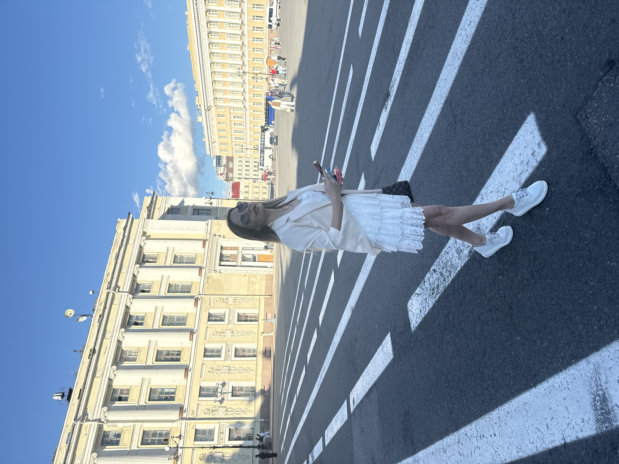 A woman stands on a pedestrian crossing in Palace Square, Saint Petersburg, Russia. She is wearing a white lace dress and a light-colored blazer, with white sneakers. She holds two mobile phones in her hands. The square is bordered by grand, neoclassical buildings, characteristic of Saint Petersburg's architecture, bathed in sunlight. In the background, other people and vehicles are visible, suggesting a bustling urban environment. The sky is blue with scattered clouds.