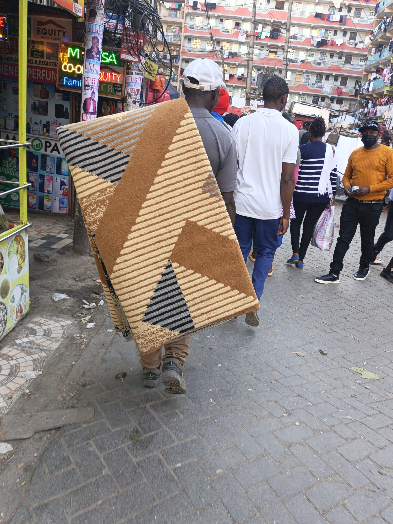 The image depicts a crowded street scene in Nairobi, Kenya. The main subject is a man carrying a large, folded rug or carpet on his shoulder. The rug features a geometric pattern with brown, tan, and blue-gray stripes and triangular shapes. The man is wearing a white baseball cap and a gray shirt.
Several other individuals are walking on the street, including a man in a white shirt and blue jeans, a woman in a striped shirt, and a man in an orange sweater and black pants wearing a face mask. The street is paved with gray interlocking stones.
In the background, there are several multi-story buildings with clotheslines strung between them, laden with laundry. Various commercial signs and advertisements are visible on the storefronts, including signs for "Equity Bank," "Co-op Bank," and a perfume store. A food cart is visible on the side of the street. Power lines and other utility wires are strung overhead.