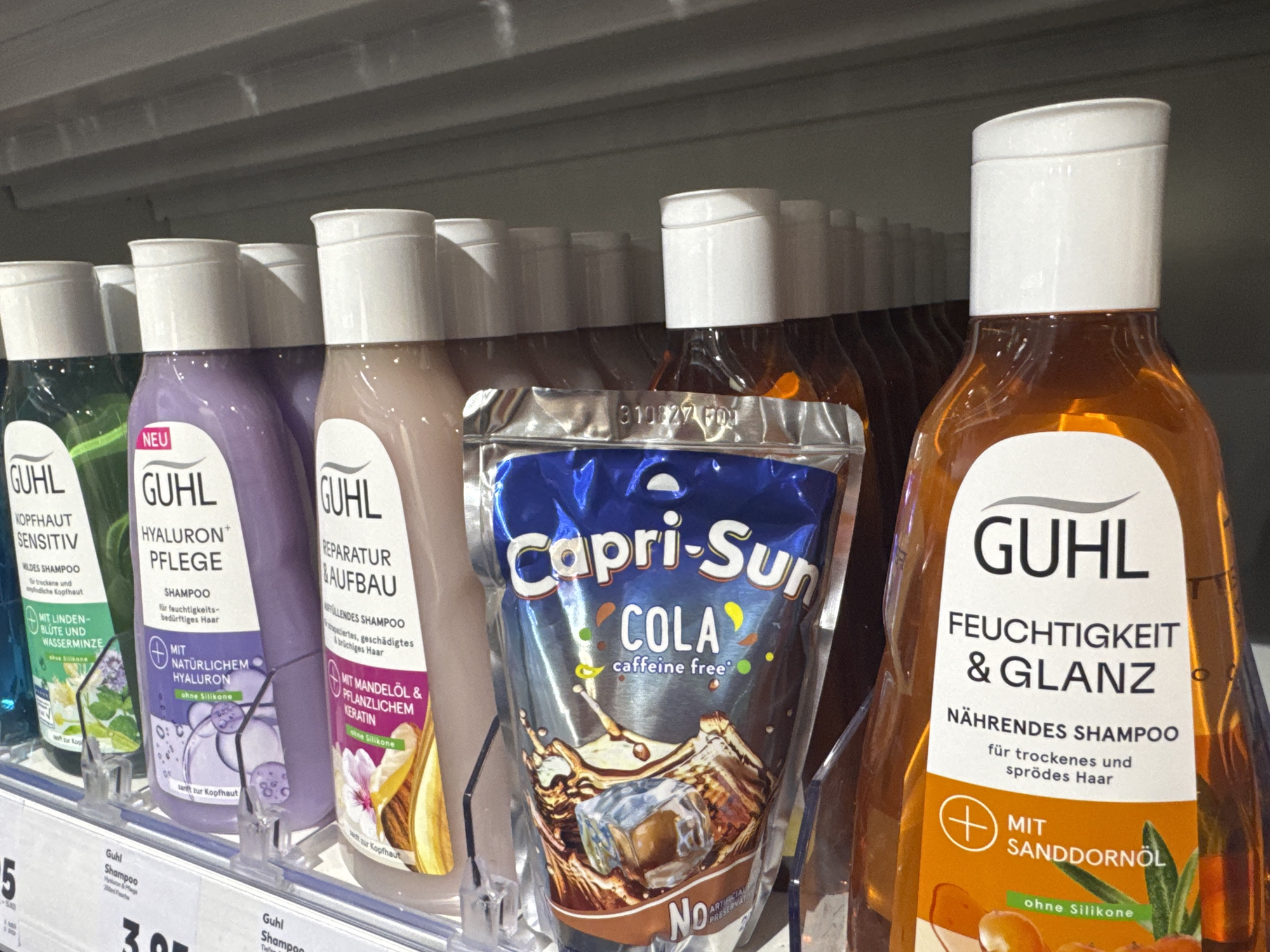 The image shows a retail shelf in a store in Waldshut, Germany, displaying various hair care products and a drink pouch. In the foreground, from left to right, are several bottles of GUHL shampoo. The first bottle is green and labeled "KOPFHAUT SENSITIV" (Scalp Sensitive). Next is a purple bottle of GUHL shampoo labeled "HYALURON PFLEGE" (Hyaluron Care). To its right is a beige bottle of GUHL shampoo for "REPARATUR & AUFBAU" (Repair & Build). In front of these shampoo bottles is a blue pouch of Capri-Sun Cola, described as "caffeine free". To the far right is an orange bottle of GUHL shampoo labeled "FEUCHTIGKEIT & GLANZ" (Moisture & Shine) and "NÄHRENDES SHAMPOO" (Nourishing Shampoo), with "MIT SANDDORNÖL" (With Sea Buckthorn Oil) prominently featured. The products are arranged on a white shelf with clear plastic price tag holders. The background shows more shelves with out-of-focus products. A price tag reading "3.95" is visible on a lower shelf, indicating prices in Euros. The lighting suggests an indoor retail environment. There are no people visible in the image.
