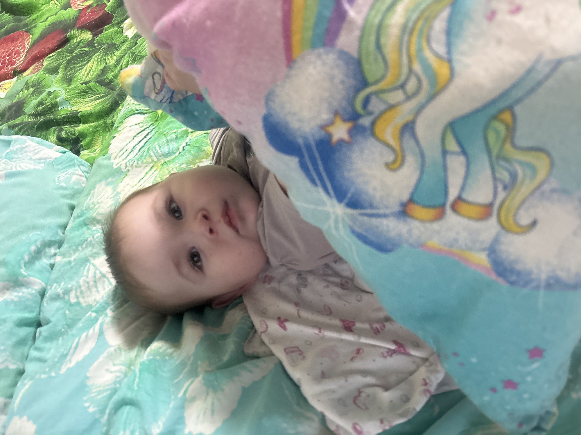 An infant is lying on its back, positioned slightly off-center to the left of the frame. The infant appears to be under one year of age, with light brown hair visible on the scalp, dark, wide-set eyes, and a neutral expression while looking directly towards the camera. Small, reddish blemishes are discernible on both cheeks. The infant is wearing a light-colored, long-sleeved garment, possibly light gray or off-white, and is partially covered by a white blanket or sleep sack featuring faint pink/purple abstract patterns, including what appears to be the letter "J" and other small shapes.
The infant is surrounded by multiple blankets and bedding. Behind the infant, to the left, is a light teal or mint green blanket with a white floral or leafy pattern. To the upper right, another blanket or sheet features a vibrant green foliage pattern interspersed with red, strawberry-like fruit elements. In the foreground, primarily covering the infant's lower body and extending to the right, is a light blue blanket featuring a large, stylized white unicorn with a blue, yellow, and green mane and tail. A rainbow motif is partially visible behind the unicorn's head, and scattered pink stars are present on the blanket's surface. A pink blanket or portion of the unicorn blanket is also visible on the infant's right side. The lighting appears soft and even, suggesting an indoor setting. The image is captured from a slightly elevated perspective looking down at the infant.