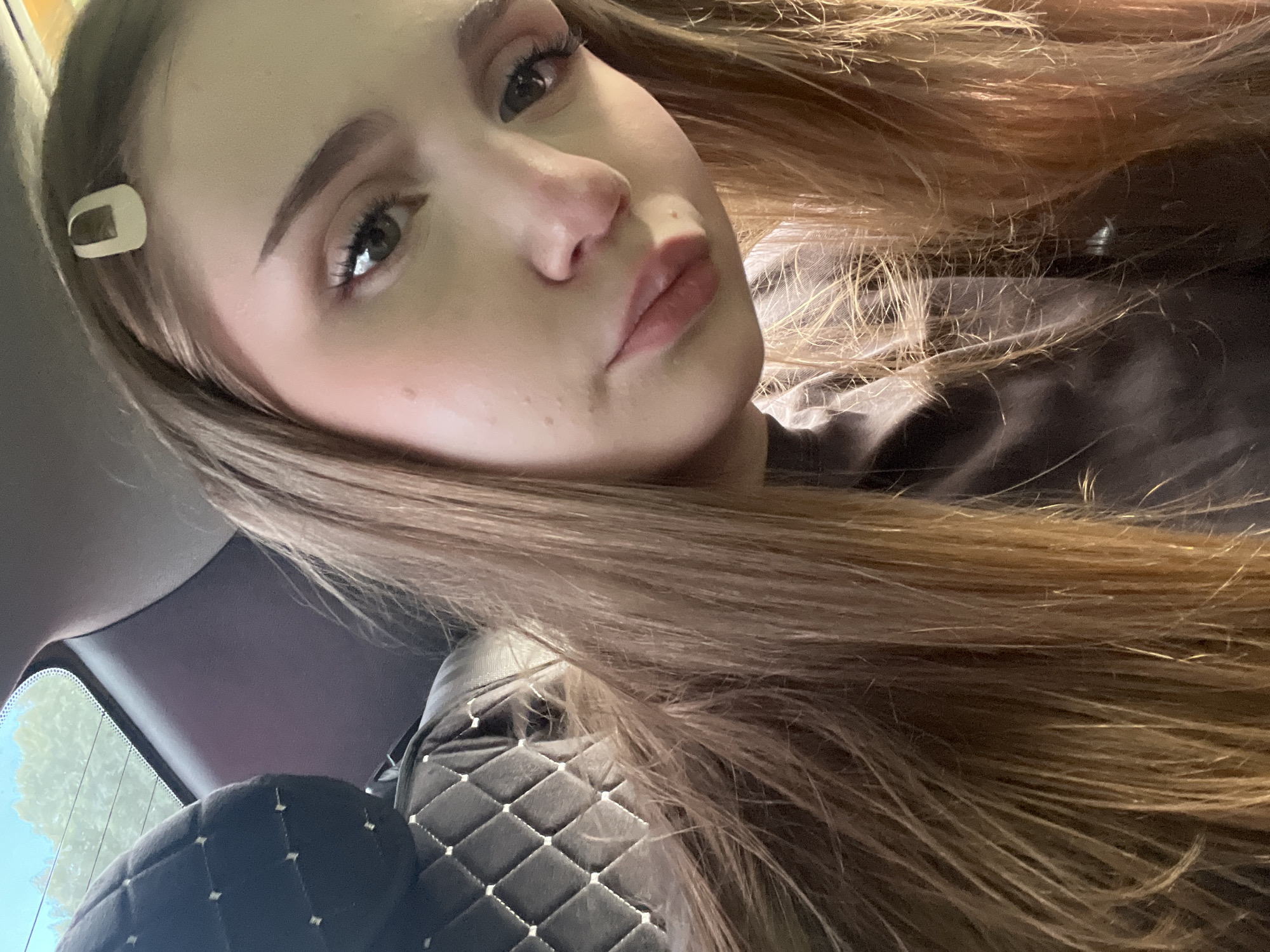A close-up, eye-level, medium shot captures a young woman with light brown hair, styled with a light-colored barrette, looking directly at the camera. Her face is lightly made up with blush and eyeliner. She is seated in the interior of a vehicle, as indicated by the quilted headrest and window frame visible on the left side of the frame. The background outside the window appears to be foliage and sky, suggesting an outdoor setting. The lighting is somewhat dim, possibly indicating it is daytime. The specific location in Novaya Otradovka, Russia, cannot be determined from the visual information provided.