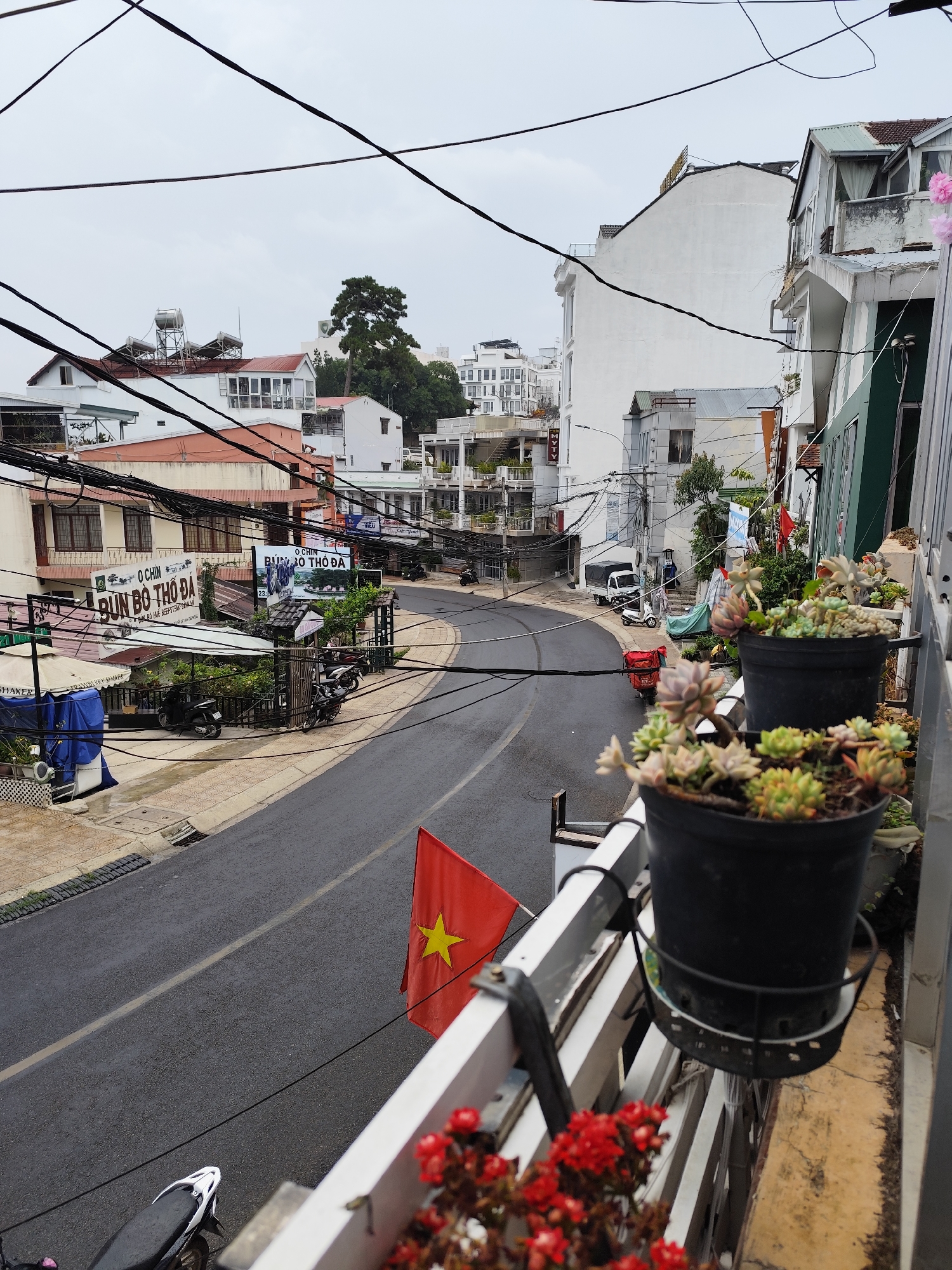 A street scene in Da Lat, Vietnam, observed from an elevated perspective, reveals a narrow asphalt road curving gently between buildings. A Vietnamese flag hangs from a balcony. Power lines crisscross overhead. Restaurant signs advertise "Bun Bo Tho Da" (a beef noodle soup), which can be seen on the left of the image. A red motorbike with a delivery bag drives up the road. Vegetation including succulents and red flowers grow in pots on a balcony railing. Other motorbikes park near the restaurants. A person stands near a white van further up the road. The weather appears overcast.