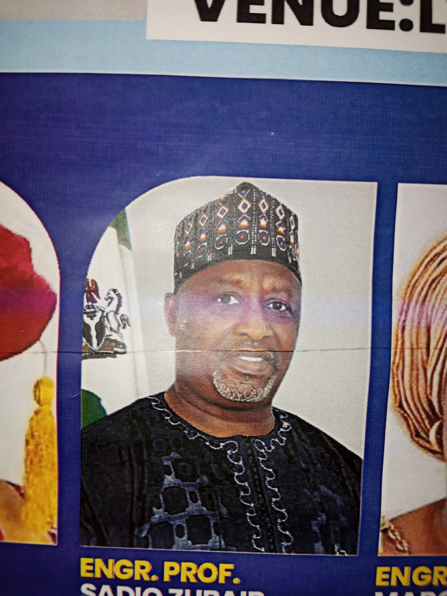 The media file is a photograph of a poster featuring individuals, likely for an event or directory. The main subject is a man with dark skin, wearing a black cap with a geometric pattern and a black embroidered garment. He is smiling and looking towards the camera. Behind him, a Nigerian flag is visible. To the right of this man, a portion of another person's head and shoulders is visible, with hair styled in distinct stripes. To the left, a blurred image of a red object and a yellow tassel can be seen.
The poster has text at the top, partially visible, reading "VENUE: L". Below the central image of the man, there is more text that is mostly legible: "ENGR. PROF. SADIO ZUBAIR". To the right of this, another section of text begins with "ENGR". The overall background of the poster is a dark blue textured material. The lighting appears to be artificial, and the image quality suggests it was taken indoors or in a controlled environment, possibly a public notice board or a printed program. There are no clear indicators of time of day, weather, or specific emotions beyond a pleasant expression from the main subject. The context provided suggests this is likely related to an event or organization in Jalingo, Nigeria.