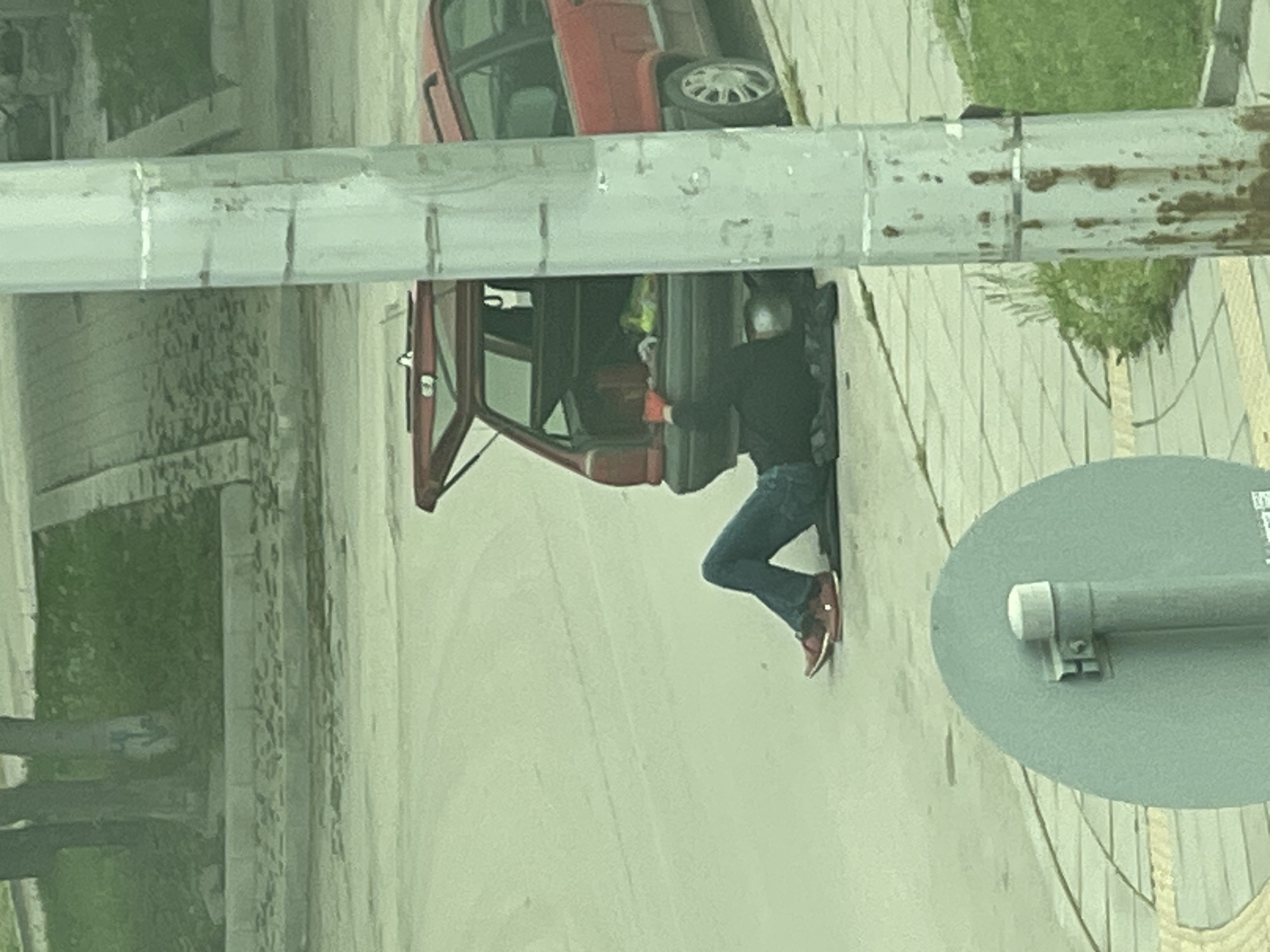 A high-angle shot depicts a red car with an open door parked on a paved surface, next to a sidewalk bordered by greenery and a low wall. A person wearing blue jeans, a black top, and red shoes is standing near the open car door. The environment features street furniture, including a circular gray sign with an attached metal post. The location is in Sofia, Bulgaria.
