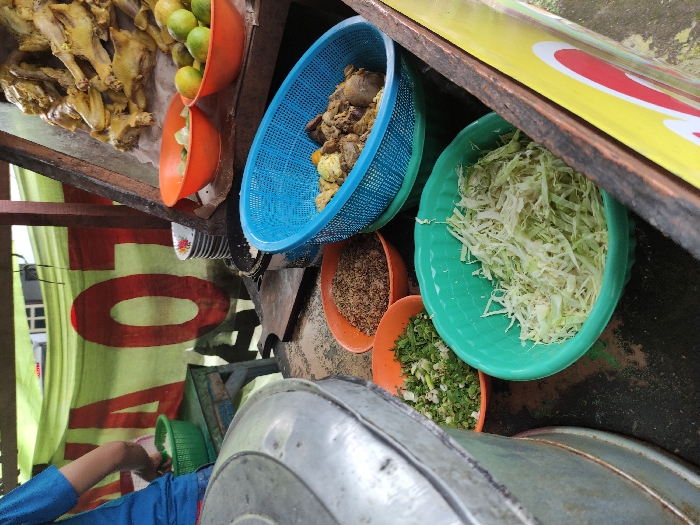 The scene is a food stall in Kecamatan Tulangan, Indonesia. It is located at latitude: -7.473, longitude: 112.632. The stall is made of wood and has a yellow and red sign. A person is preparing food. There is a large metal pot, a green bowl of chopped greens, and a green bowl of shredded cabbage. There is also a blue bowl with chicken, and an orange bowl with chicken. There are also several other bowls and dishes on the stall. There are pieces of chicken in the top right corner of the frame. The surface of the stall is wood. The sign on the stall appears to read "TOYA." The person appears to be putting food in the large metal pot. There is a sign on the wall behind the person. The person is wearing a blue shirt and appears to be facing to the left. The background is blurry and there are other objects in the scene, but they are out of focus.