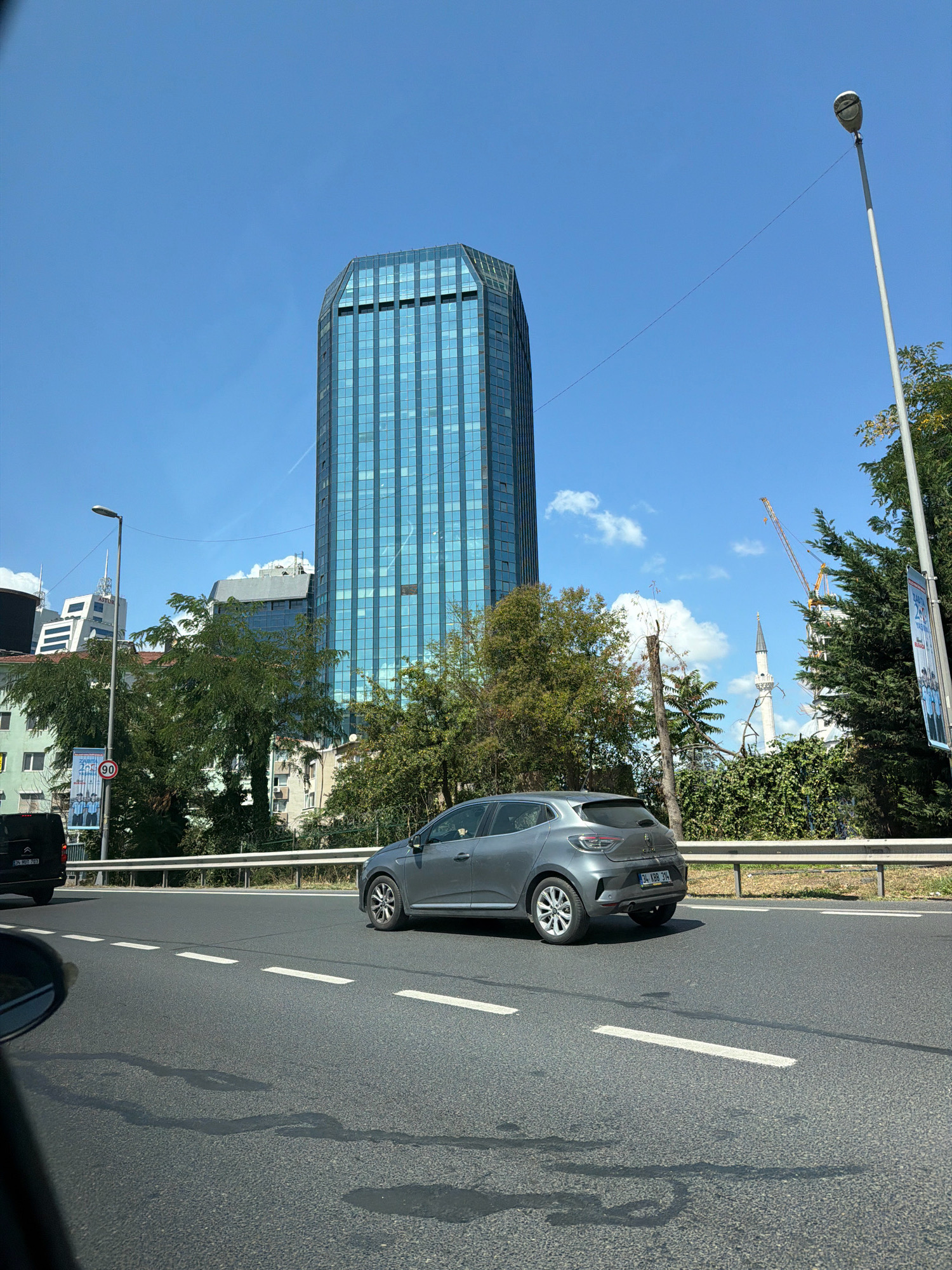 A low-angle, eye-level, full shot captures a modern, glass-facade skyscraper dominating the skyline under a clear blue sky with scattered white clouds. The building features a distinctive octagonal shape. In the foreground, a gray car drives on a multi-lane asphalt road, indicated by white lane markings and a wet patch on the asphalt. To the right of the car, a silver metal guardrail separates the road from a lush green area with trees and foliage. Behind the trees, a minaret of a mosque with a pointed spire is visible, along with construction cranes. A street light stands tall on the left side of the frame. A black van is partially visible in the far left of the image. A billboard with a red circle and a "90" inside is also present. The overall scene suggests a sunny day in an urban environment, likely Şişli, Turkey, given the architectural style and presence of a mosque.