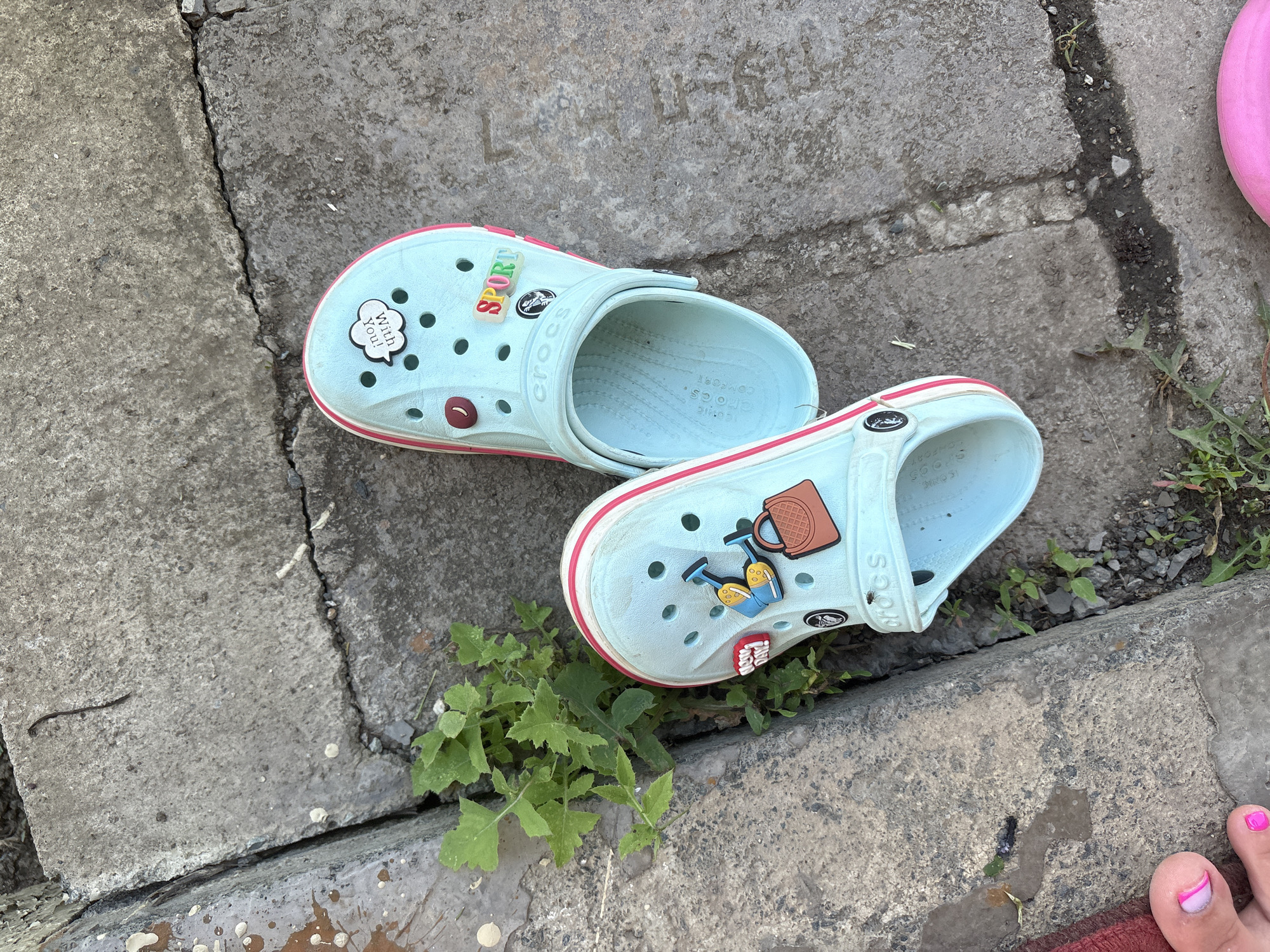 A pair of light blue Crocs with pink trim are lying on a cracked concrete sidewalk in Orsk, Russia. One Croc has several Jibbitz charms including a speech bubble that says "With you!", a red button, a brown handbag, and two yellow watering cans. The other Croc has a "SPORT" Jibbitz and another Jibbitz of a black and white logo. Green weeds are growing through the cracks in the sidewalk, and a small fly is visible on the right shoe. In the bottom left corner, a person's toes with bright pink nail polish are visible. A red edge is also visible in the bottom left corner. The scene is outdoors, likely on a sunny day.
