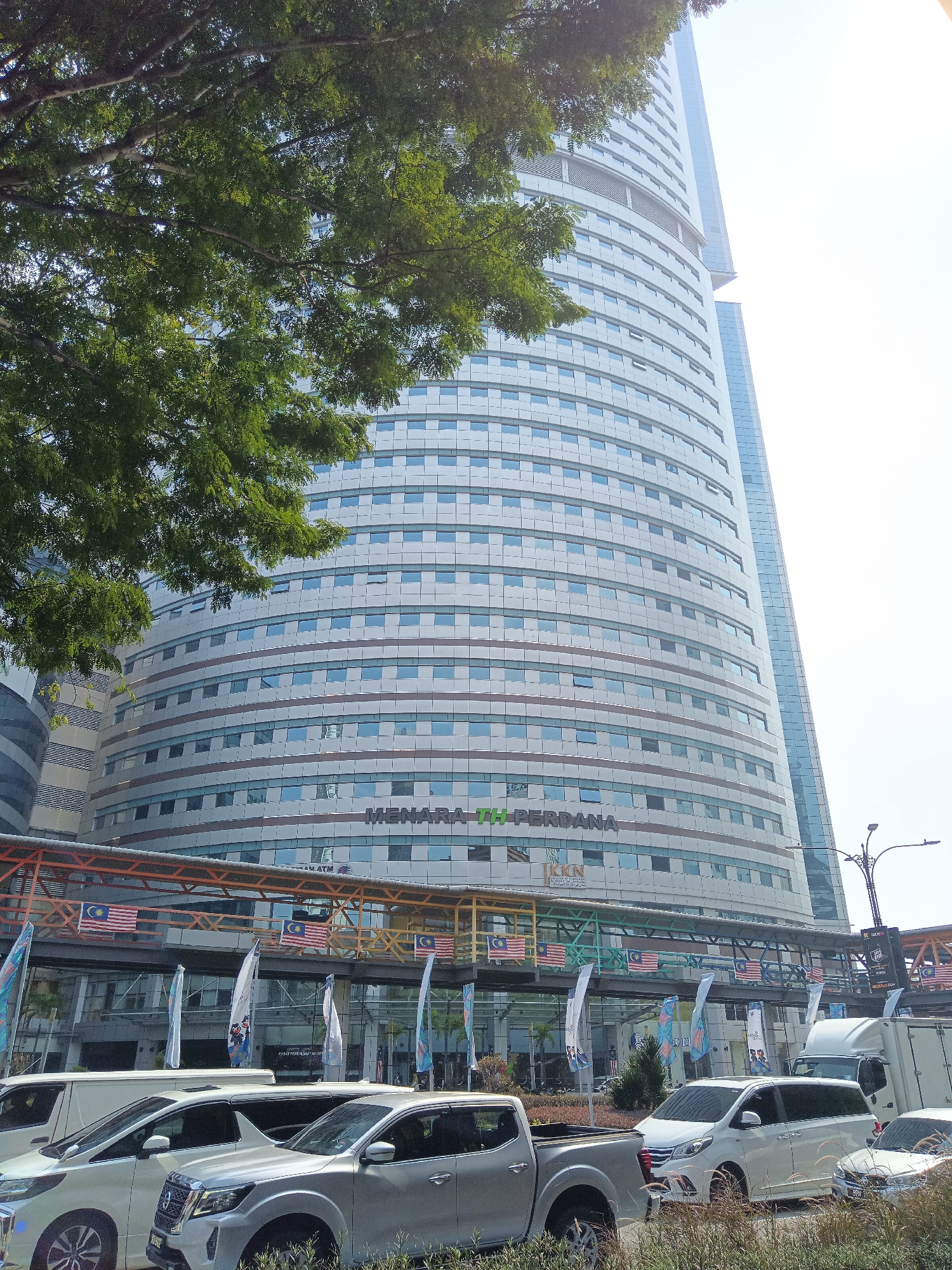 In the city of Kuala Lumpur, Malaysia, a curved, modern skyscraper with a light-colored facade and numerous windows dominates the frame. The skyscraper is flanked by a taller, glass-paneled building to its right. In the foreground, several white vehicles, including a pickup truck, a van, and a car, are parked on a street. A walkway with Malaysian flags draped across it connects the lower floors of the skyscraper to an adjacent structure. Lush green foliage from a tree partially obscures the upper left portion of the skyscraper. The sky is bright and appears to be overcast.