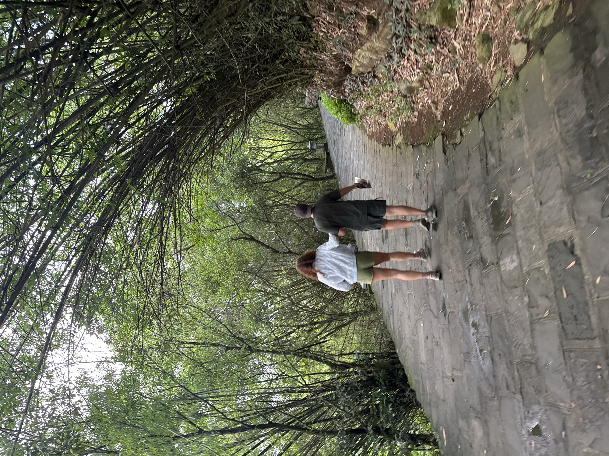 Two people, a woman and a man, are walking away from the camera on a stone pathway lined by dense bamboo and other greenery. The woman is on the left, wearing a light-colored jacket and olive-green shorts. The man is on the right, wearing a dark t-shirt and dark shorts, and he is holding a folded piece of paper or pamphlet. The pathway is wet, suggesting recent rain. The scene appears to be a park or garden path within the city of Tây Hoa Lư Ward, Vietnam, characterized by lush vegetation and a natural, somewhat overgrown environment. The path curves to the right, leading further into the bamboo grove.