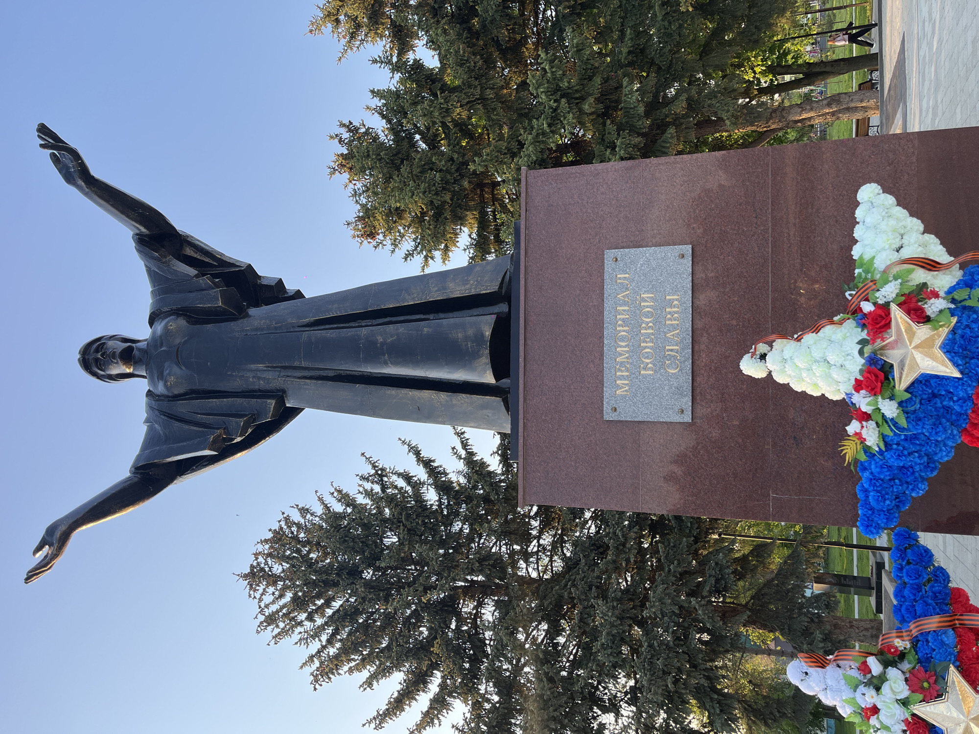 The image shows a monument dedicated to war heroes, located in Novopokrovskaya, Russia. A bronze statue of a woman with outstretched arms stands beside a reddish-brown rectangular pedestal. A stone plaque with text in Russian is attached to the pedestal. A wreath of white, red, and blue flowers is attached to the pedestal with the Ribbon of Saint George. A gold star is also visible on the wreath. Evergreen trees stand behind the monument. The sky is clear and blue.