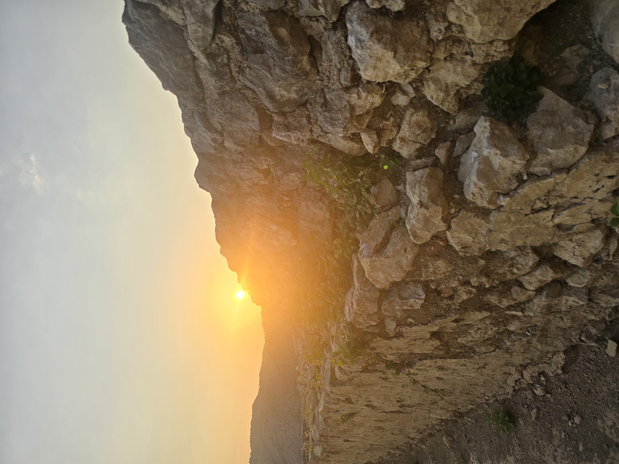 The image shows a stone wall under a bright orange sunset in Gornja Vrbica, Montenegro. The wall is made of rough, uneven stones and has small green plants growing between them. The sun is low in the sky, casting a warm glow over the scene and creating lens flares. In the background, there are rolling hills. The overall impression is one of natural beauty and a sense of history.