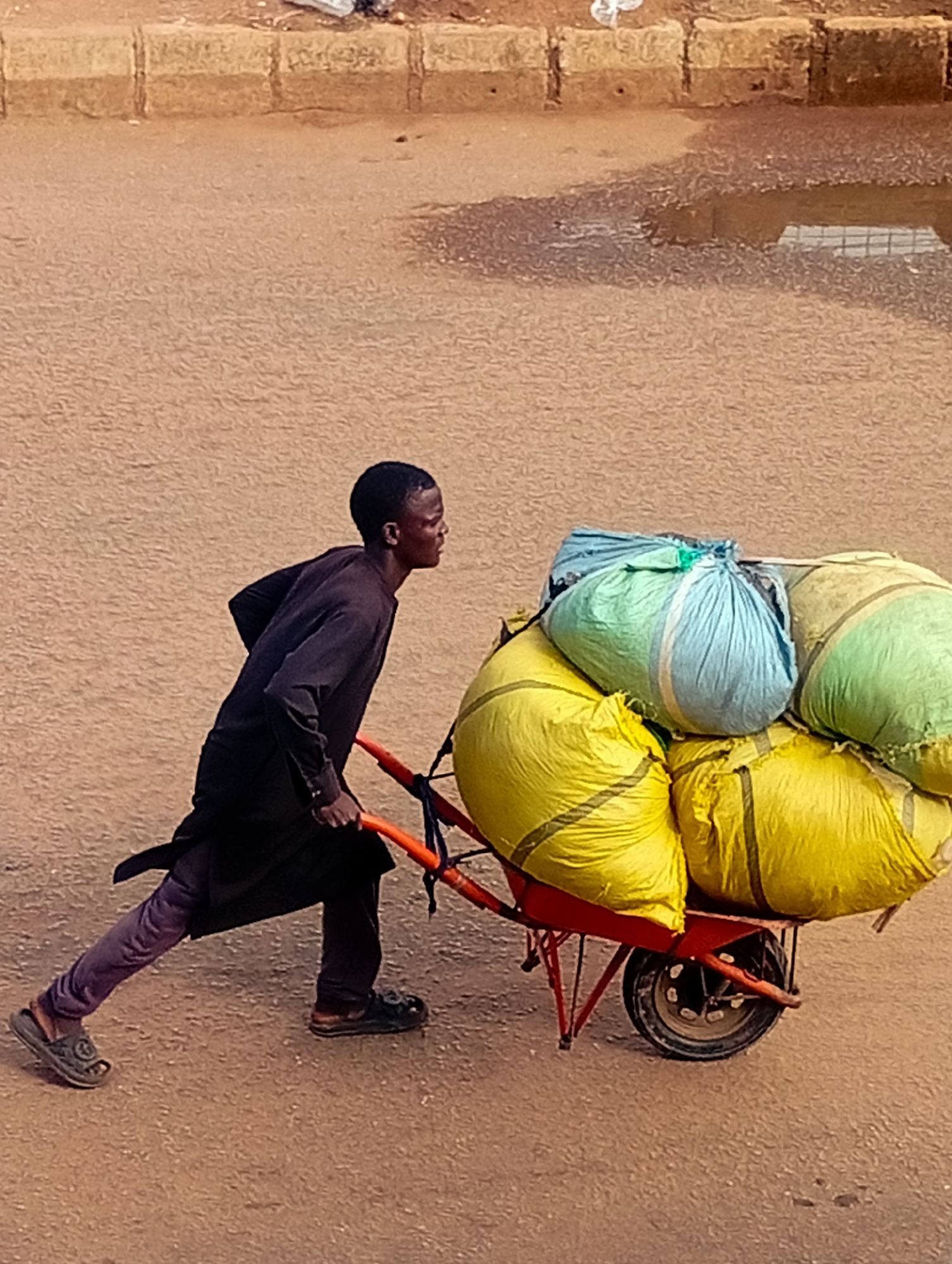 A young Black male is pushing a red wheelbarrow loaded with several large, brightly colored bags. He is wearing a dark, long-sleeved garment and purple leggings. The wheelbarrow is laden with at least three yellow bags, one light blue bag, and one green bag. The ground is a dirt or gravel surface, and there are puddles of water visible in the background. The scene appears to be in an outdoor setting, possibly a street or pathway. This image was taken in Bukuru, Nigeria.