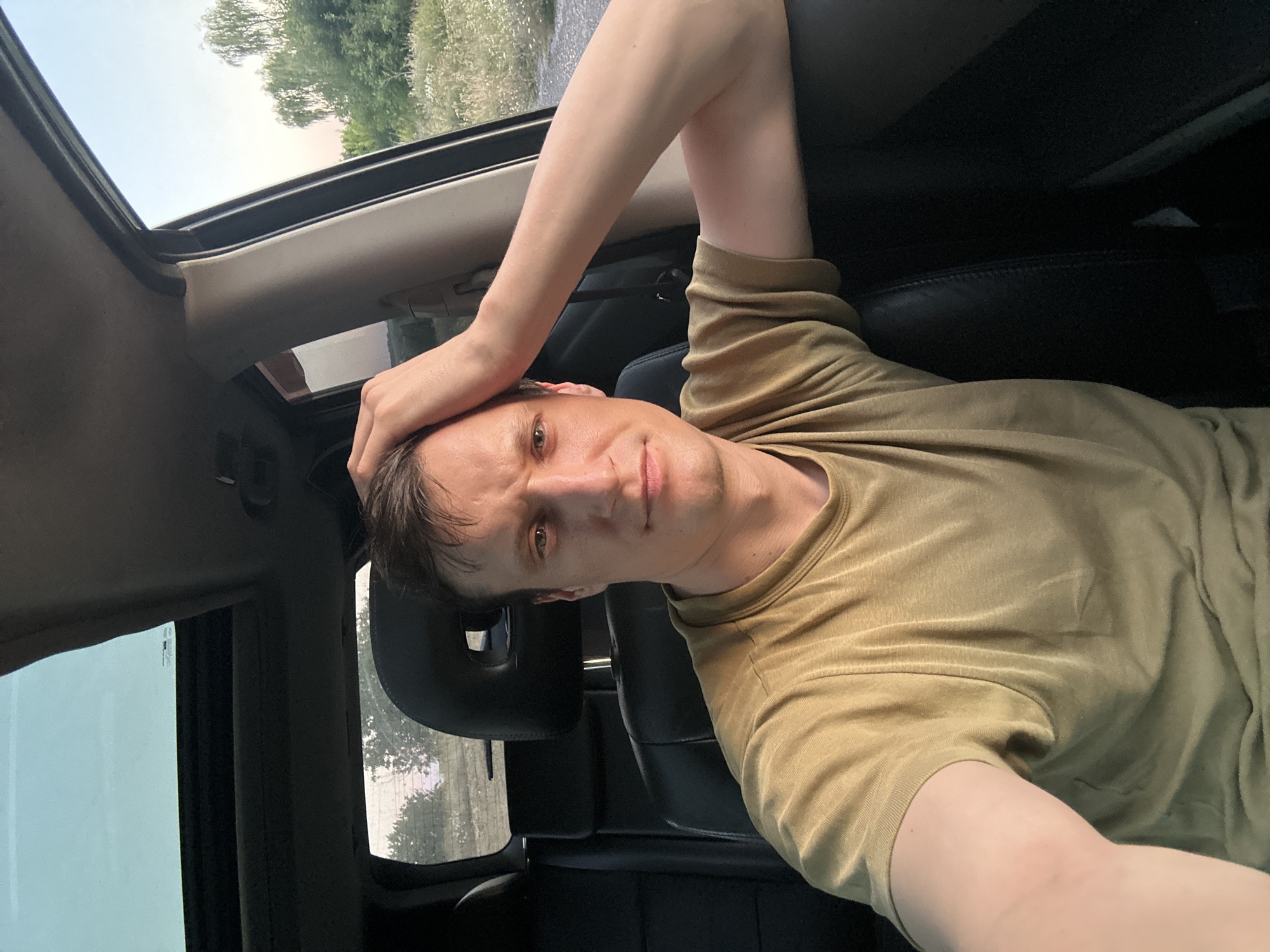 A man is sitting in the driver's seat of a vehicle, looking at the camera. He is wearing a light brown t-shirt and has his right hand resting on the back of his head. The vehicle has a sunroof that is open, allowing daylight to enter. The interior of the car appears to be black leather. Outside the car, trees and vegetation are visible, suggesting a rural or suburban setting. The time of day appears to be late afternoon or early evening, given the warm lighting. There are no visible landmarks in the image that would indicate the specific location of Makiv, Ukraine.