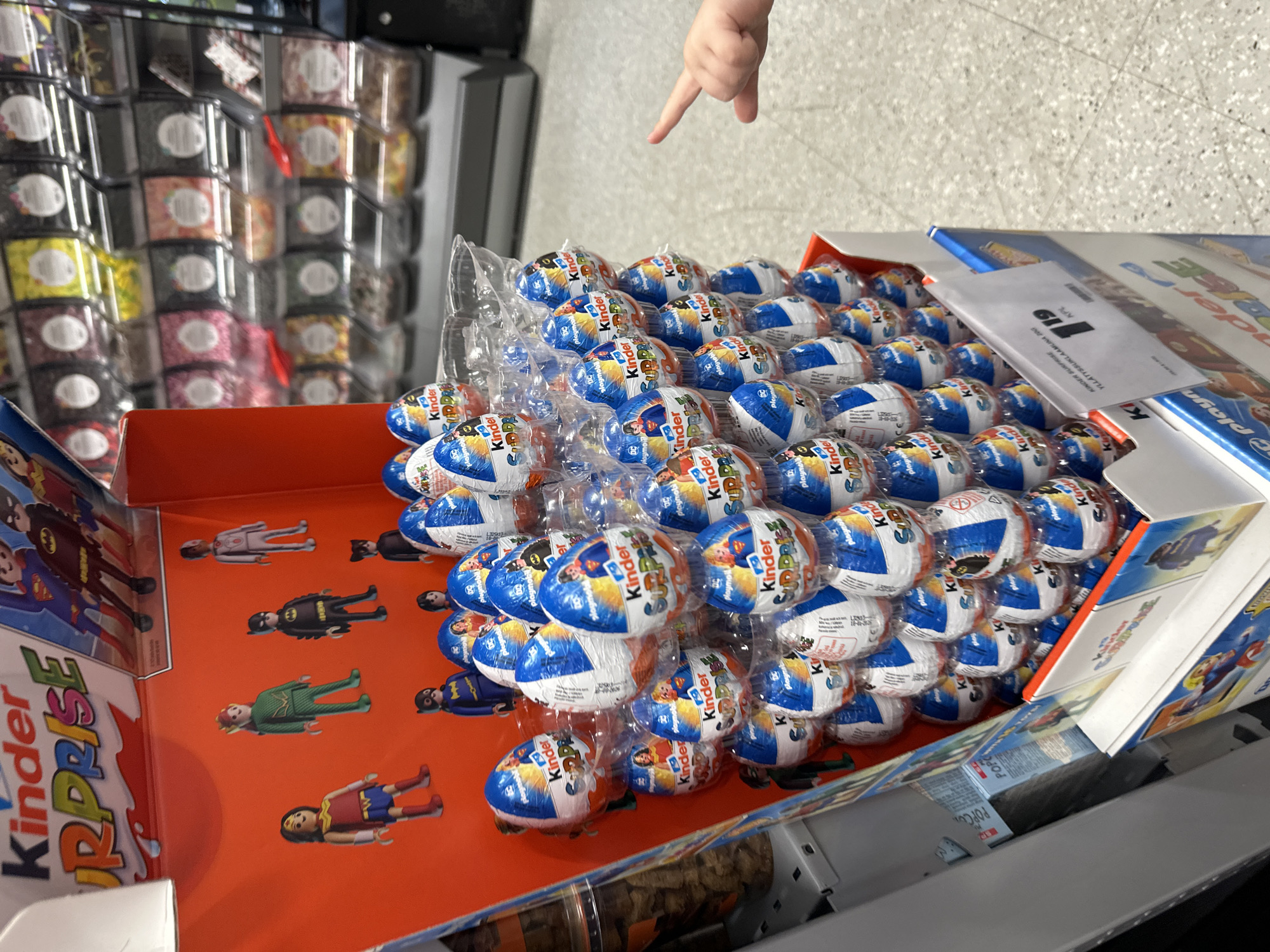 An indoor shot captures a point-of-purchase display of Kinder Surprise eggs in a retail setting. The display is primarily orange and features various superhero characters from DC Comics and Playmobil figures. The eggs are stacked in clear plastic packaging, with most of them wrapped in the signature foil of Kinder Surprise. The background shows shelves stocked with bulk candies, displayed in clear bins. A young child's hand, with a pointed index finger, is visible in the lower right corner of the frame, reaching towards the display. A price tag indicating "1 19" is attached to the display. The floor appears to be a speckled tile.