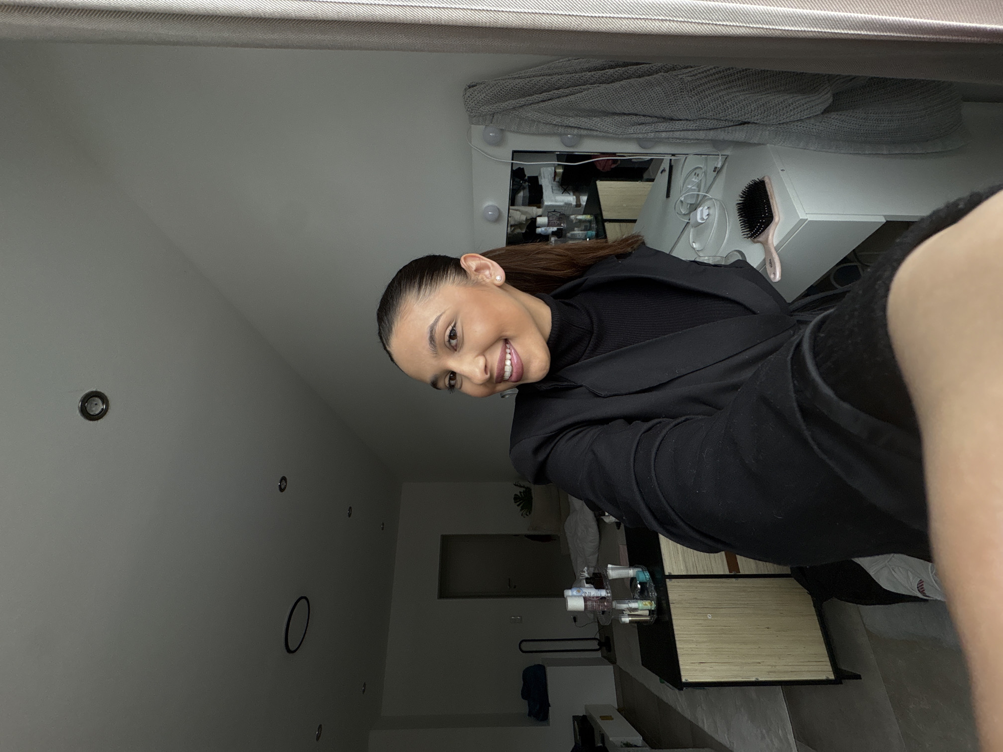 A young woman with brown hair pulled back in a ponytail smiles at the camera. She is wearing a black turtleneck and a black blazer. The background includes a vanity with a mirror, lights, and various beauty products, as well as a glimpse of a room with a light-colored door and some furniture. The location is possibly a residential interior, potentially in Mönchengladbach, Germany.
