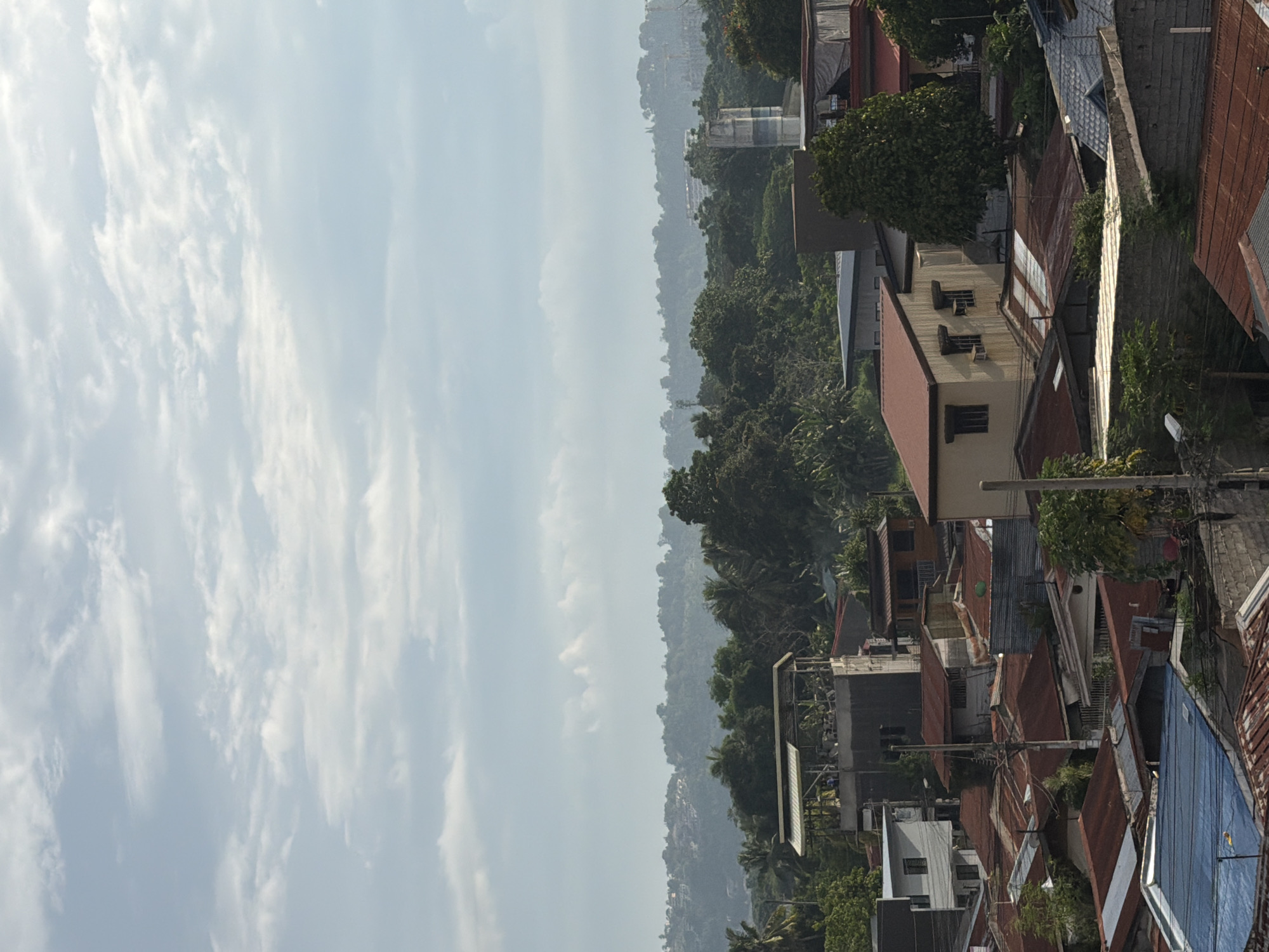 The image shows an elevated view over a section of Davao City, Philippines. The foreground features densely packed residential buildings with predominantly red corrugated iron roofing. Electrical lines are visible, weaving among the buildings. Beyond the built environment, a lush, green hillside rises, fading into a hazy sky. The sky is filled with white, streaky clouds, suggesting potentially humid weather conditions.