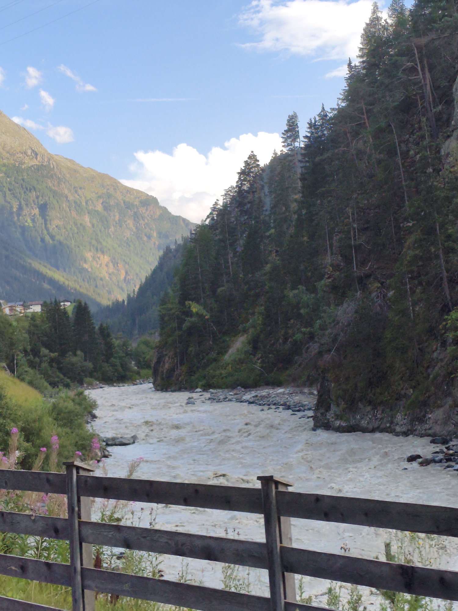 The image depicts a pastoral scene in the Oetz Valley, near Längenfeld, Austria. A wide, muddy river flows through the center of the frame, its churning waters suggesting a strong current. On the right bank, a steep, densely wooded mountainside rises, covered in coniferous trees that cast long shadows. The left side of the image features a softer, gentler slope covered in green grass and sparse vegetation, with a few buildings visible in the distance nestled amongst the trees. In the foreground, a rustic wooden fence made of horizontal planks and vertical posts runs across the width of the image, partially obscuring the view of the river and the wildflowers growing alongside it. The sky above is a bright blue, dotted with fluffy white clouds. The overall impression is one of natural beauty and tranquility, with a hint of untamed wilderness in the dramatic mountain landscape.