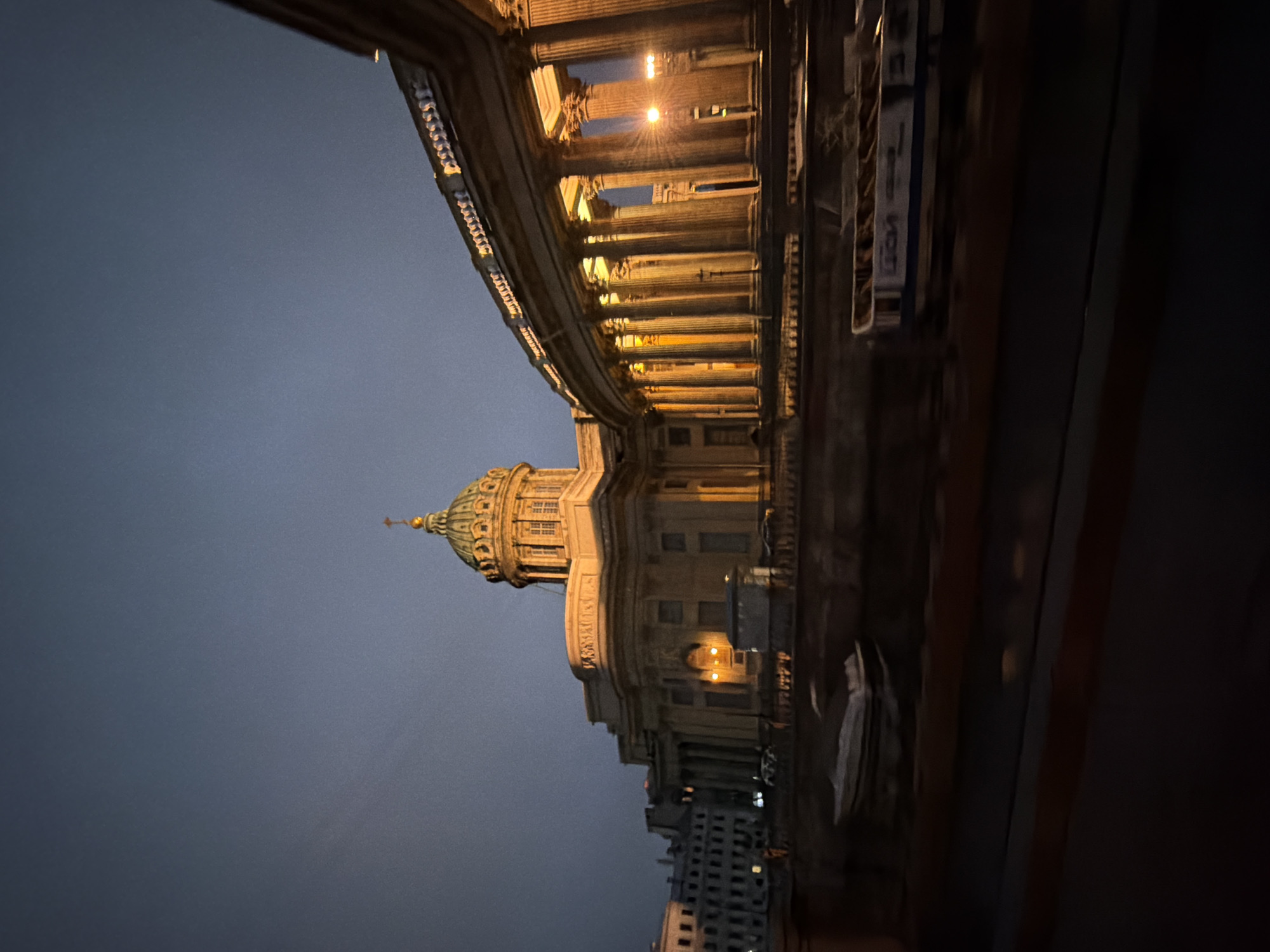 The image shows the Kazan Cathedral in Saint Petersburg, Russia, at dusk. The grand cathedral, with its iconic colonnade and dome, is illuminated by warm artificial lights, casting a golden glow on the neoclassical architecture. The sky is a deep twilight blue, with hints of grey clouds. In the foreground, a dark body of water, likely a canal, reflects the lights. Several boats are visible on the water, one of which appears to be a tour boat. The overall atmosphere is serene and majestic, highlighting the architectural beauty of the landmark.
