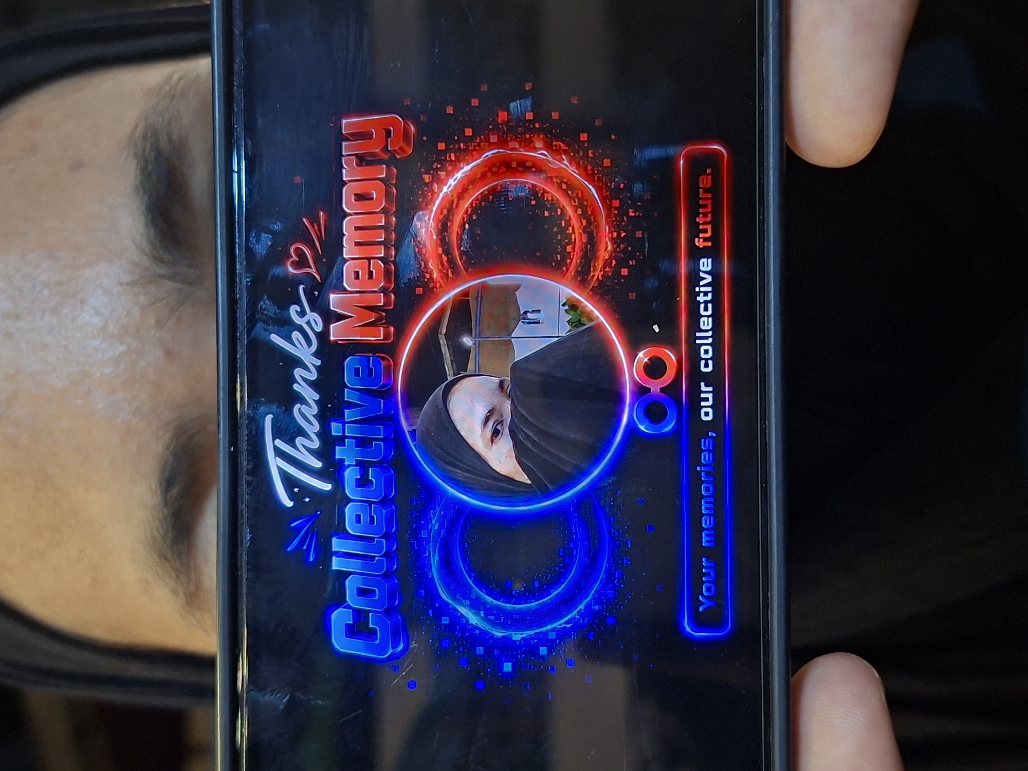 A person is holding a smartphone, displaying an image with the text "Thanks Collective Memory. Your memories, our collective future." A circular image within the display shows a person wearing a hijab. Red and blue animated effects surround the text and the circular image. A person's face and fingers are partially visible on either side of the phone, suggesting they are holding the device. No location in Lhokseumawe, Indonesia is evident in the image.