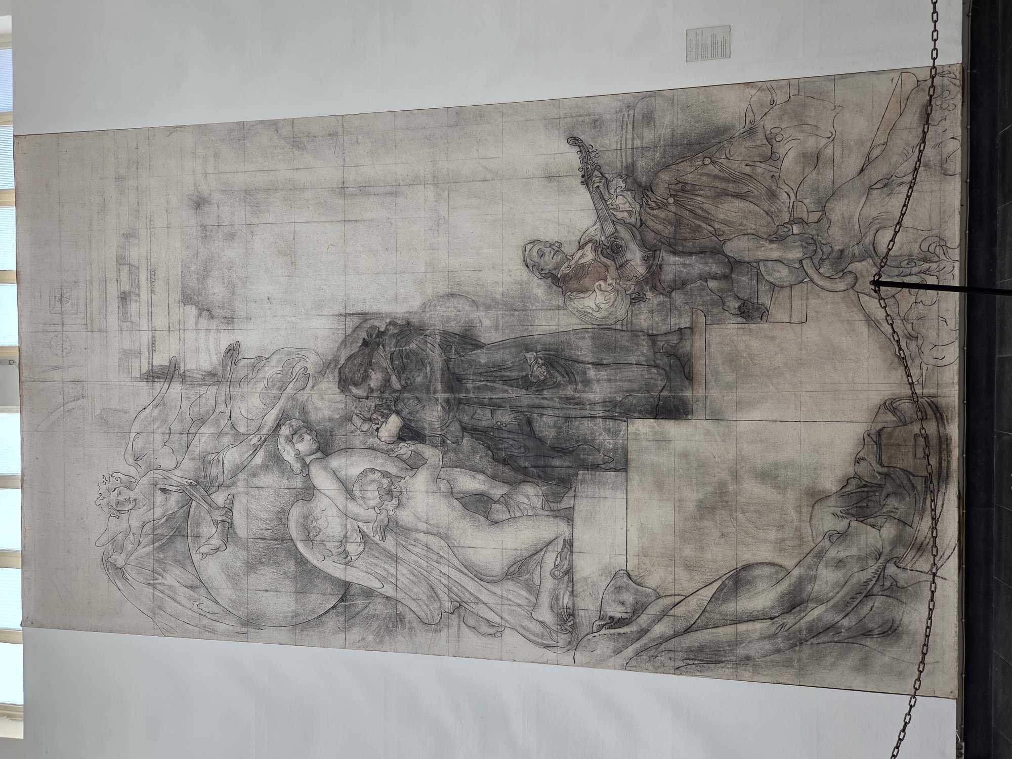 This is a detailed sketch on a large canvas, depicting a complex allegorical scene with multiple figures. In the upper section, a winged figure, possibly an angel, is seated on a globe, with another winged figure reaching up towards it. Below them, a third figure, partially draped, is kneeling. To the right of these figures, a robed individual is standing and interacting with a seated musician playing a lute-like instrument. The lower portion of the sketch shows draped fabric and other indistinct forms. The sketch is rendered in shades of gray and black with visible grid lines, indicating it may be a preparatory study for a larger mural. A small plaque is visible on the right wall. The artwork is displayed in the Malmö City Hall, located in Stortorget, Malmö, Sweden.