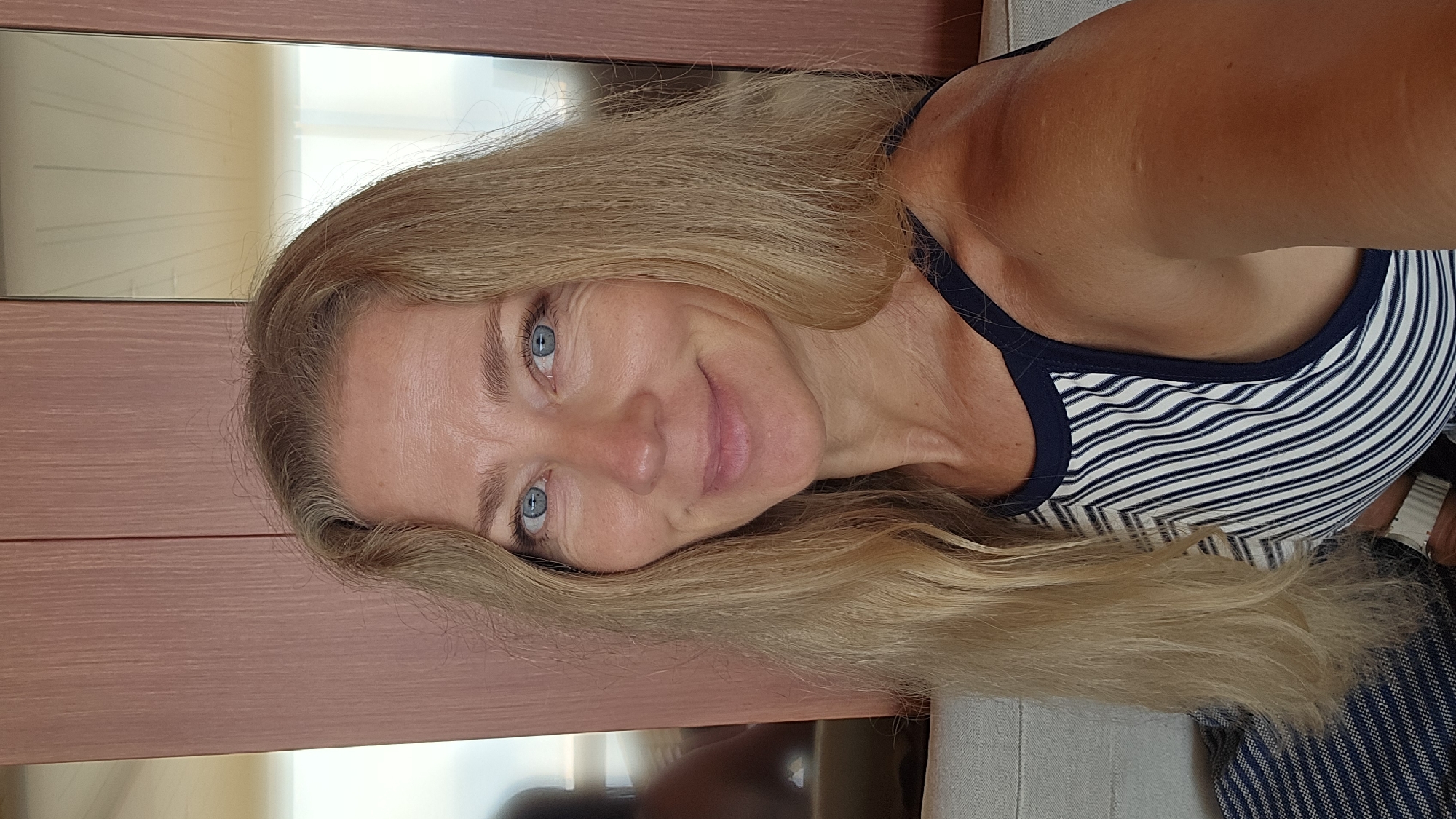 A middle-aged woman with long, blonde hair and blue eyes is smiling in a selfie. She is wearing a striped tank top with a dark blue trim. The background appears to be an interior with a light pinkish-tan wooden wall behind her head and a glimpse of a mirror. The setting does not provide definitive information for identification of a location in Barcelona, Spain.