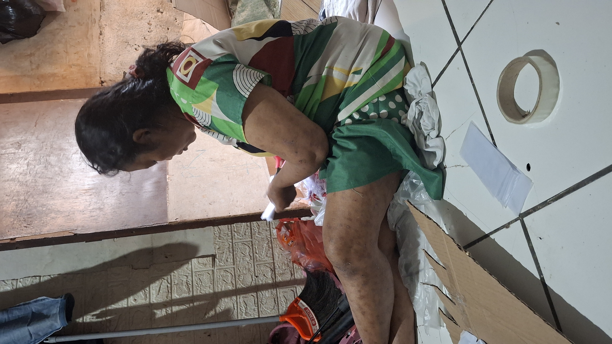 A person sits on the floor in a dimly lit room, holding a small white object, possibly a piece of paper or a phone, and looking down at it. The person is wearing a colorful, patterned shirt and a green skirt. Their legs are visible and appear to have skin conditions. Scattered around the person are various objects including a broom, plastic bags, cardboard, and a roll of tape. The floor is tiled and the walls appear to be textured. The overall scene suggests a humble, indoor environment.