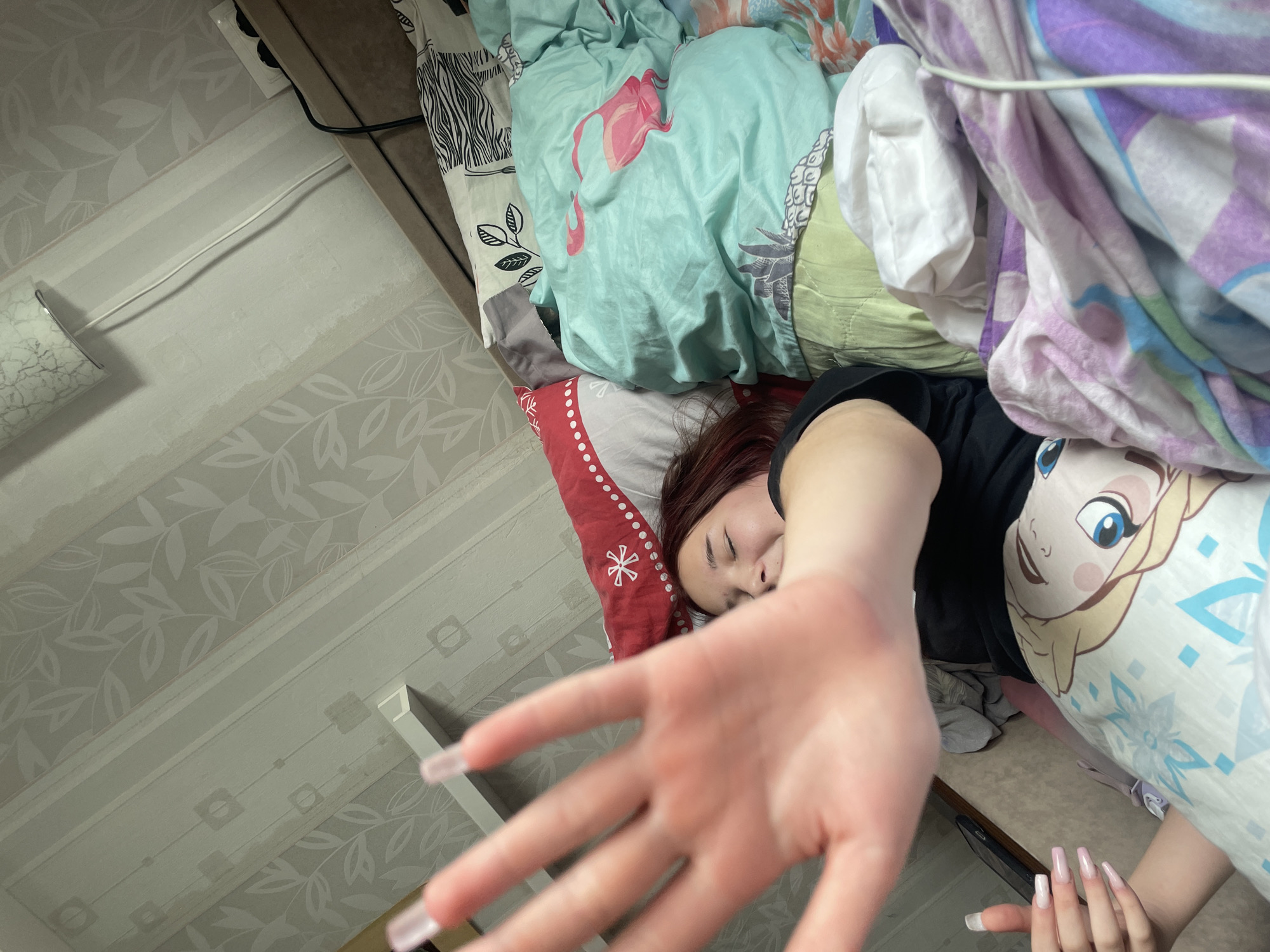 The image captures a young woman lying in bed in what appears to be a bedroom in Saint Petersburg, Russia. Her red-brown hair is visible against a red and white patterned pillow. Her eyes are closed, and a relaxed, possibly sleepy, expression is on her face. Her right hand is extended directly towards the camera, suggesting a close-up, personal moment, perhaps as she is waking up or reacting to the photo being taken. She has long, light-colored polished nails.
She is surrounded by an array of colorful and patterned bed linens, including a prominent teal blanket with pink flamingo designs, a white blanket with pineapple patterns, a purple and white plaid blanket, and a white pillowcase featuring the character Elsa from Disney's Frozen. She is wearing a black t-shirt.
The background shows the room's walls, covered in light grey wallpaper with a subtle leaf pattern. Another section of the wall features a more geometric, grid-like pattern. A white electrical cord runs along the wall, connected to an outlet near the headboard. A lamp with a textured shade is partially visible on the left side of the frame. The overall perspective is a high-angle, intimate shot. No discernible text is visible in the image.