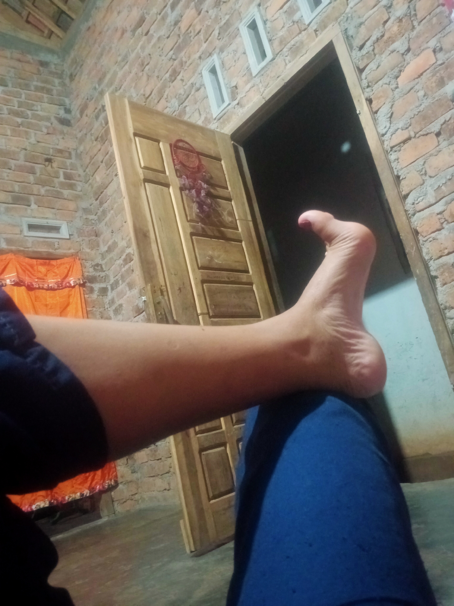 In an indoor scene of a modest home in Nusa Agung, Indonesia, a person's bare left foot and lower leg are prominently featured in the foreground, resting on what appears to be a blue cloth. The foot is extended and slightly curved, with visible toes. The leg is also visible from the knee down. Behind the person, a wooden door with paneling stands open, revealing darkness within. The walls are constructed of exposed brick. To the left of the door, an orange fabric with a decorative trim is visible, possibly curtains or a bedspread. The floor appears to be concrete. The overall lighting suggests a dimly lit interior, possibly at dusk or night.