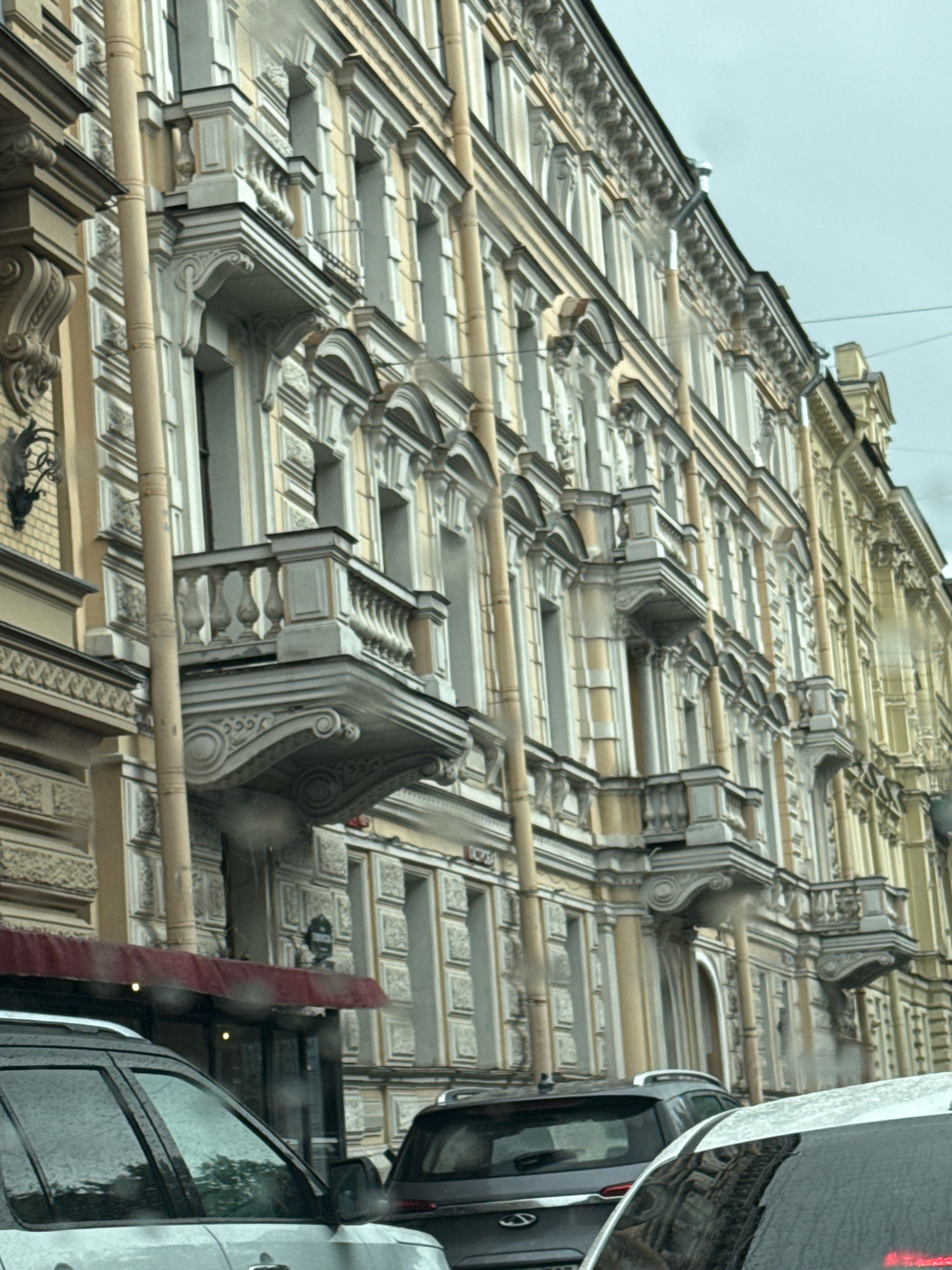 The image depicts a street scene in Saint Petersburg, Russia, focusing on the ornate architecture of a building. The facade is characterized by multiple floors with repeating window patterns and decorative balconies. Vertical beige pipes run along the building's exterior, likely for drainage. In the foreground, parts of two cars are visible, with the front portion of a light-colored SUV on the left and the rear of a darker SUV on the right. The sky is overcast, suggesting a cloudy day. The overall impression is of an urban environment with historical architectural elements.