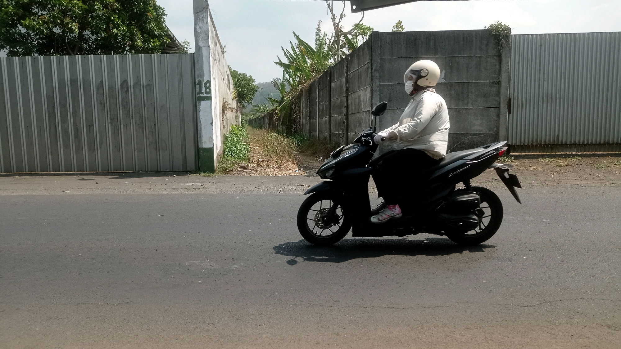 A person wearing a white helmet, face mask, and a light-colored long-sleeved shirt rides a black scooter on a paved road. The scooter has a black body and silver wheels. The person is wearing dark pants and pink and white shoes. They are holding the handlebars and their right hand is visible, wearing a white glove. The background consists of a gray concrete wall on the right and a corrugated metal fence on the left. Green vegetation, including trees and what appear to be banana plants, is visible behind the walls and fence. The sky is overcast. The scene is in Bedali, Indonesia.