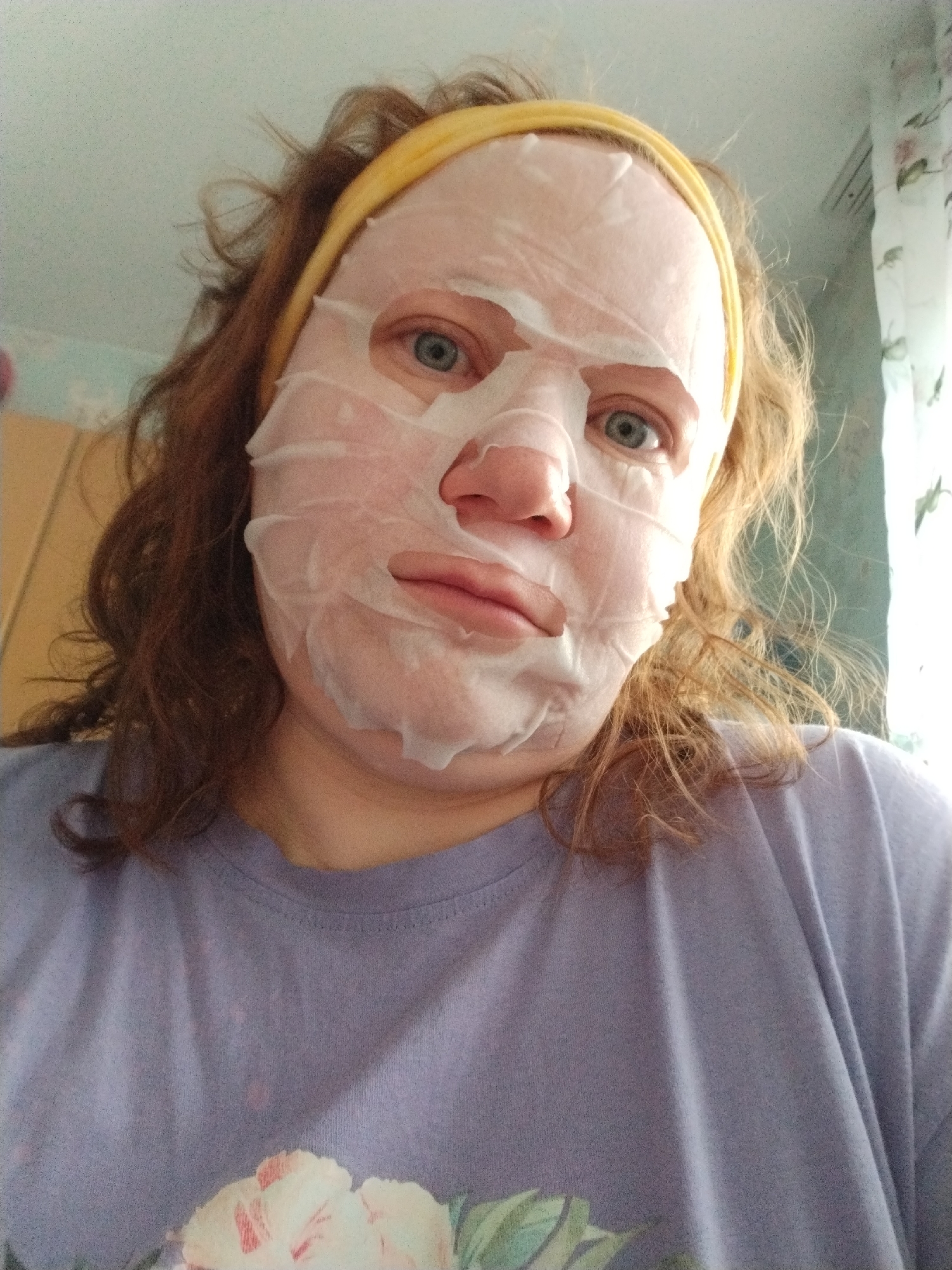 A woman is photographed close-up, likely in an indoor setting. She is wearing a sheet face mask and a yellow headband. Her hair is reddish-brown and appears somewhat disheveled. She is wearing a light purple t-shirt with a floral design near the bottom. The background is slightly out of focus, showing parts of a room with floral wallpaper, a cabinet, and a window with curtains. The image was taken in Chaikovsky, Russia.