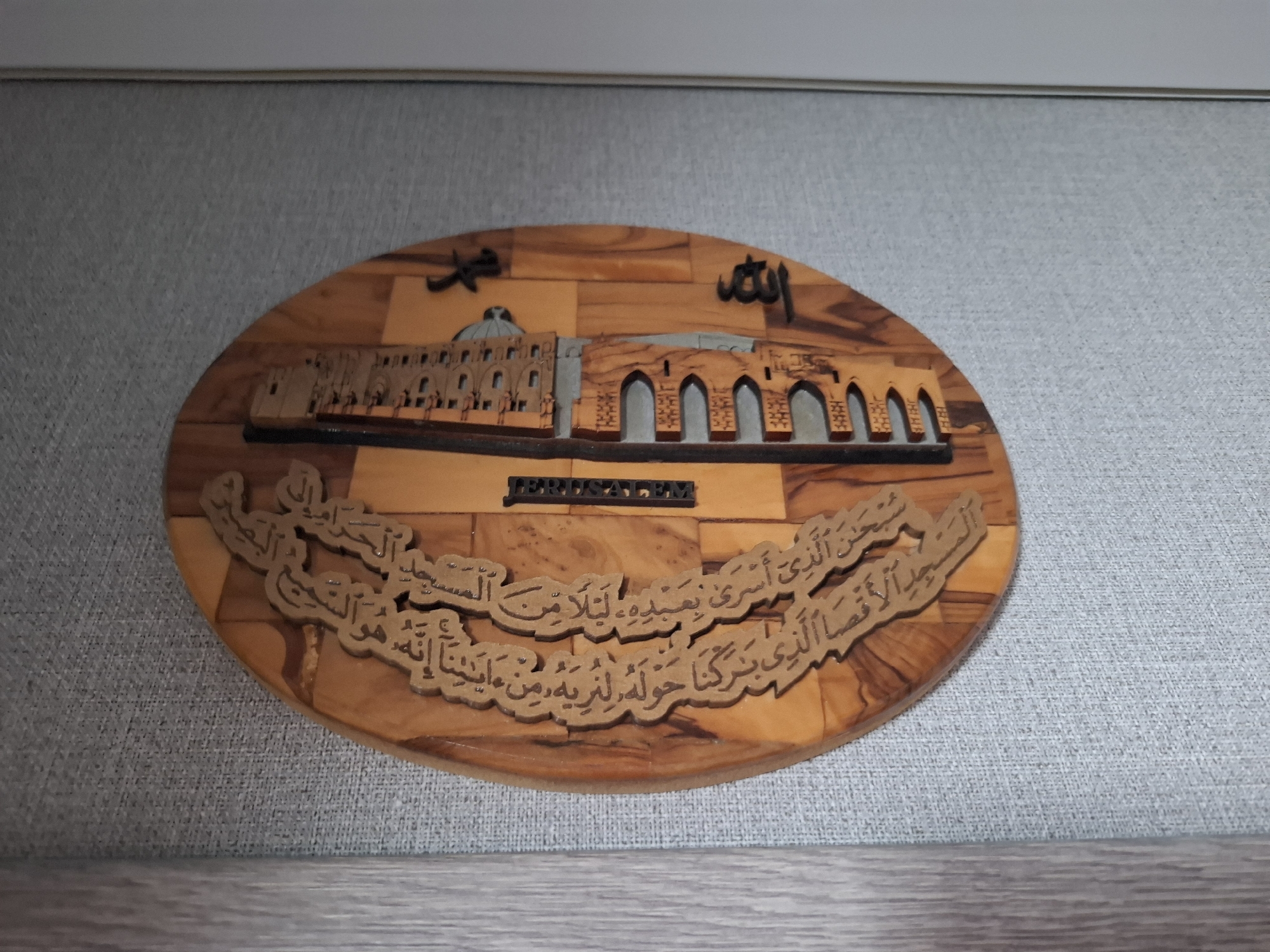 Here's a description of the image:
The image shows a wooden oval plaque mounted on a gray wall. The plaque depicts a stylized scene of Jerusalem. The Dome of the Rock and the Western Wall appear on the plaque. The word "Allah" is written in Arabic script above the buildings. Below the Jerusalem depiction, there is an inscription in Arabic. The word "JERUSALEM" is written in block letters between the buildings and the inscription. The plaque appears to be made of olive wood.