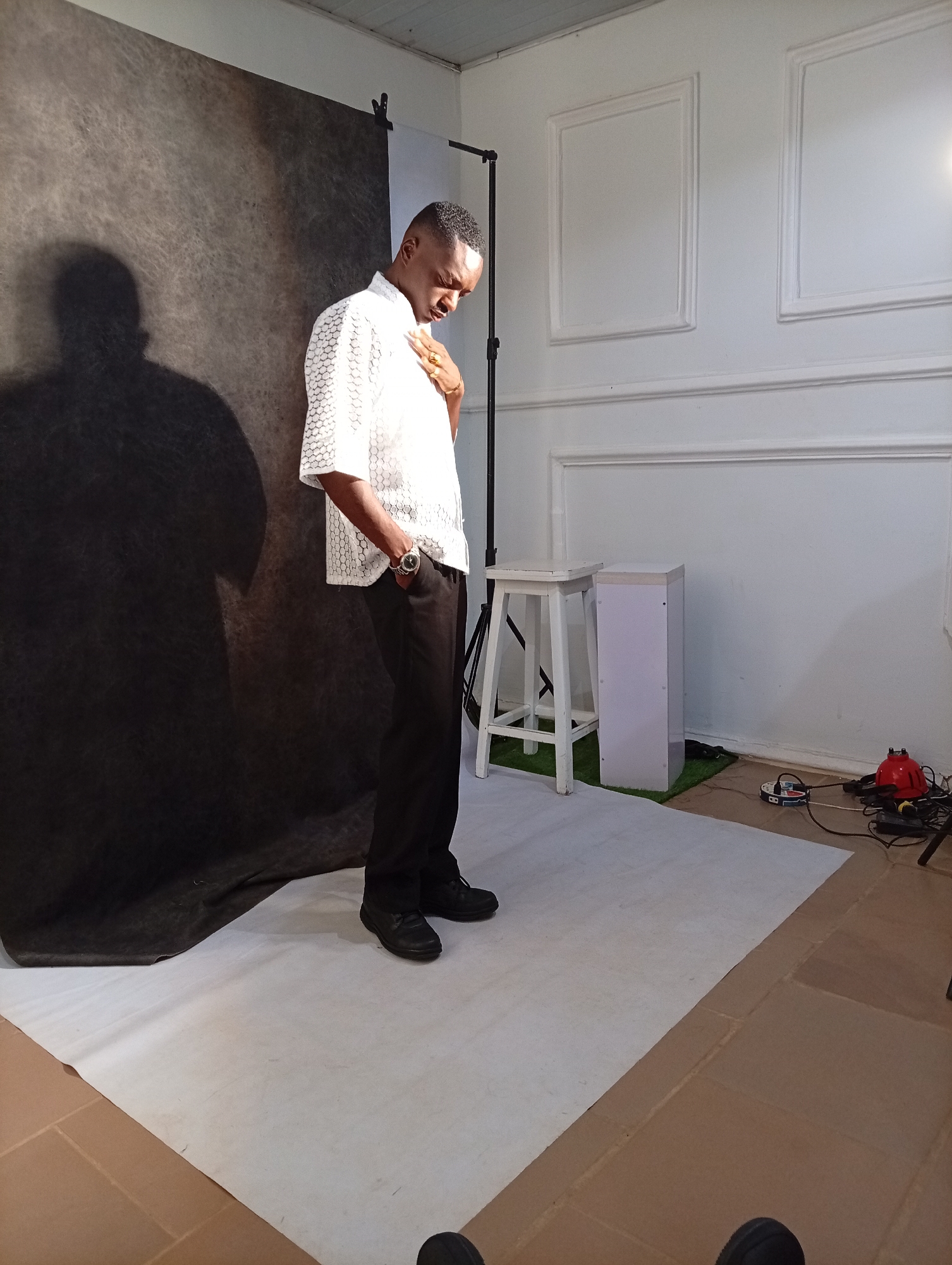 A man is standing on a white surface in a room. He has short, dark hair and is wearing a light-colored, short-sleeved shirt with a pattern, dark trousers, and black shoes. His right hand is in his pocket and he is wearing a watch on his left wrist and a ring on his right hand. He is looking down. Behind him is a gray backdrop creating a shadow. To his right is a white stool and a white cabinet against a white wall with decorative moldings. There are electrical wires and equipment on the floor to the right. A light stand is visible behind the stool. The floor is a mix of the white surface he's standing on and a brown tiled area. The location is identified as a room. The city and area remain unknown.