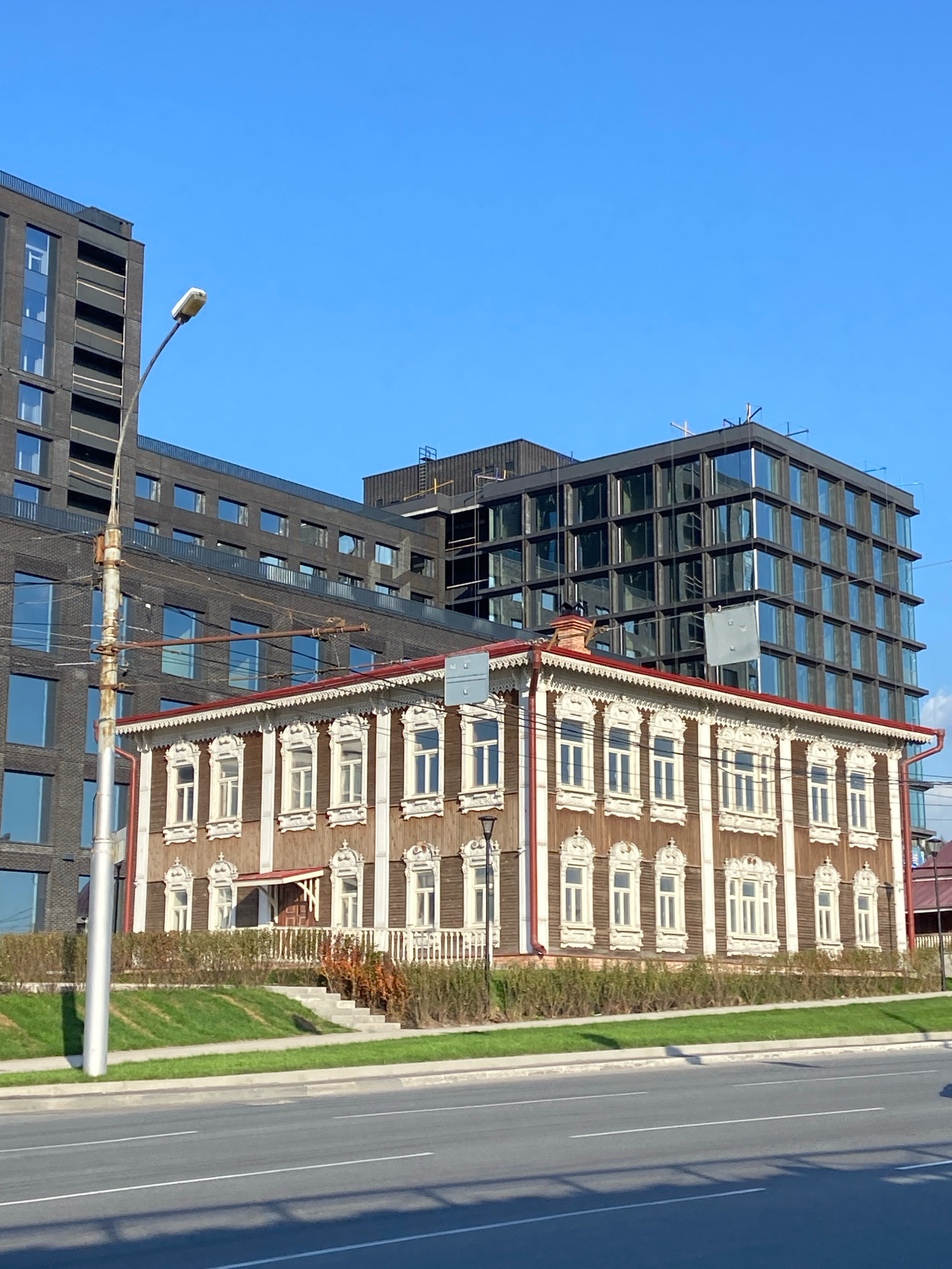 The image displays a street scene in Novosibirsk, Russia, featuring a contrast between historic and modern architecture. In the foreground, a traditional wooden house with ornate window trim and a red roof stands prominently. The house is two stories high and exhibits intricate carvings around its windows, a hallmark of old Russian wooden architecture. Behind the wooden house, a dark, modern multi-story building with numerous glass windows dominates the background. This contemporary structure appears to be an apartment complex or office building, indicating urban development. A light pole is visible on the left side of the image, with wires stretching across the scene. The area in front of the wooden house is landscaped with a green lawn and a hedge. A paved road runs across the bottom of the frame, suggesting the location is along a street or highway. The sky is clear and blue, indicating fair weather.