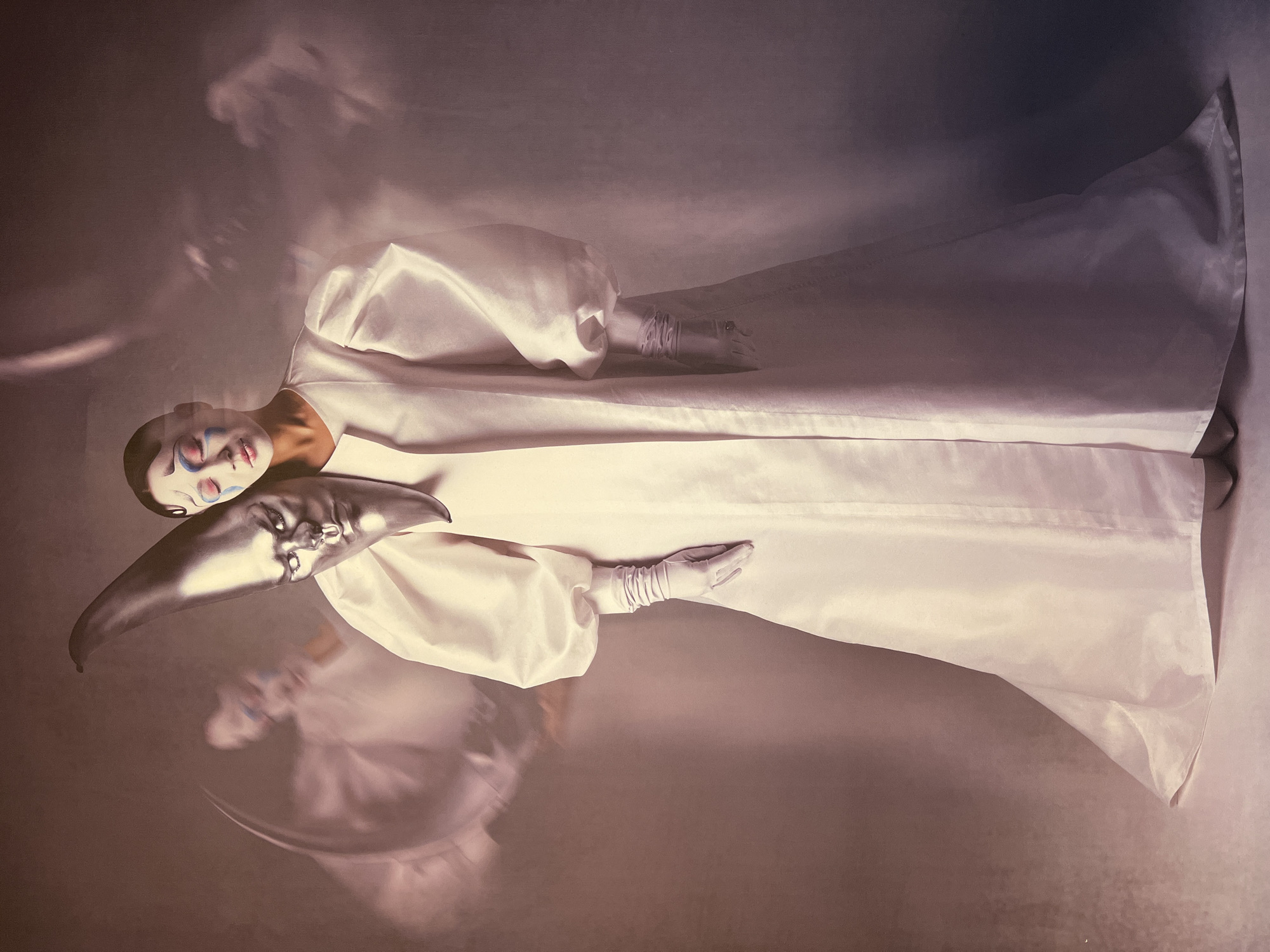 The image depicts a person with theatrical makeup, featuring blue and pink accents around the eyes and white face paint, wearing a voluminous white gown with puffed sleeves and white gloves. They are holding a silver, crescent-shaped mask with a human-like face and a nose ring in front of their own face. Ghostly, blurred figures are visible in the background, suggesting a dreamlike or ethereal atmosphere. The lighting is soft and diffused, creating a moody and dramatic effect. The image does not contain any discernible elements that would allow for its localization to the city of Vitsebsk, Belarus.