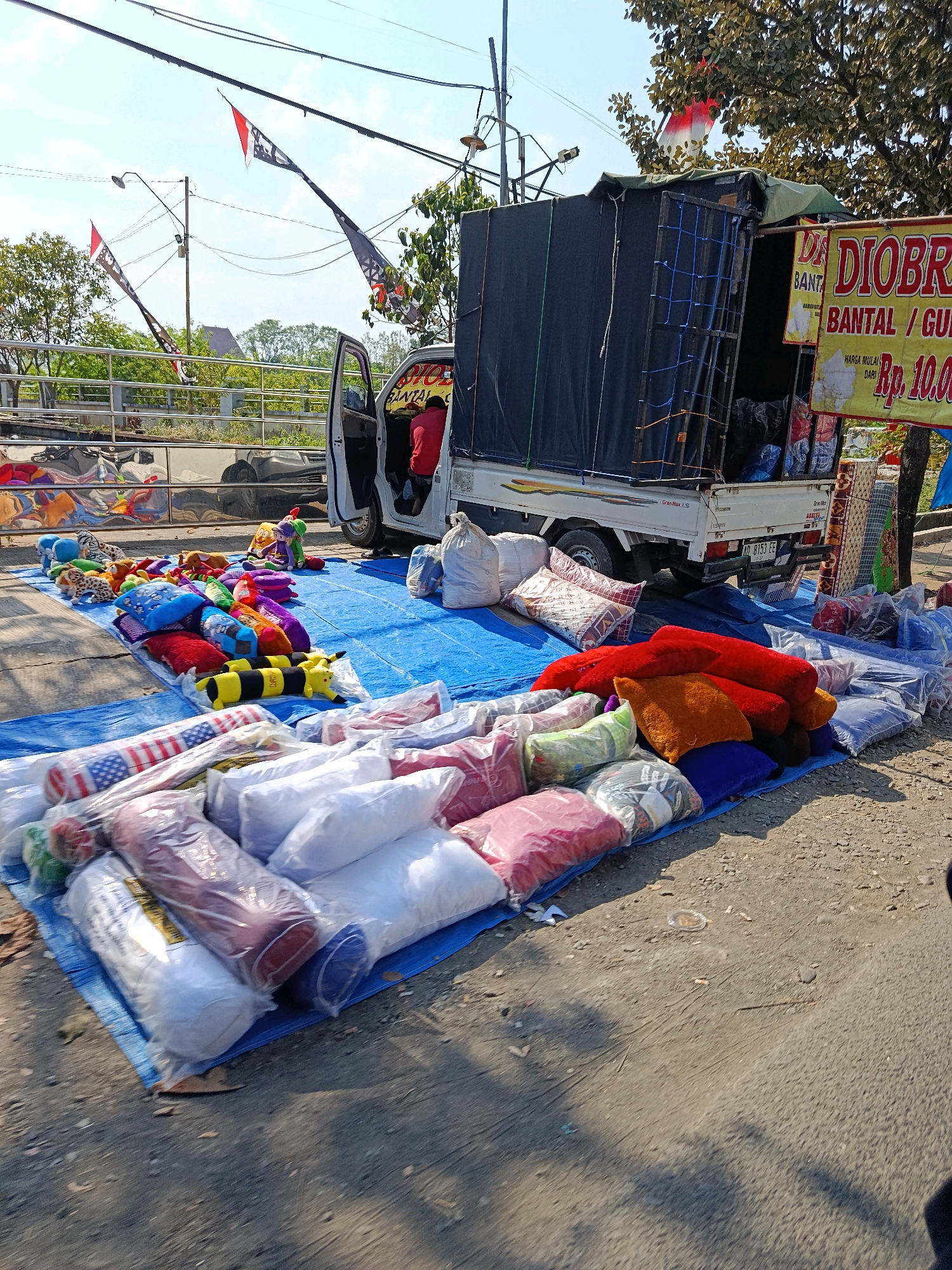 This is a full-shot, outdoor photograph taken on a sunny day in Getas Pejaten, Indonesia, featuring a roadside stall selling pillows and stuffed animals. A white pickup truck with a black tarp covering its cargo area is parked on the right side of the image, serving as the stall's base. A man in a red shirt is visible inside the truck's cabin.
In front of the truck, a large blue tarp is spread on the ground, displaying a variety of items for sale. These include numerous colorful pillows, many of which are individually packaged in plastic. Among the pillows are several stuffed animals, including a bumblebee-patterned pillow, a leopard-patterned pillow, and a purple stuffed character. Rolled items, possibly blankets or more pillows, are also visible on the tarp, some with American flag patterns.
To the right of the truck, a yellow sign with red lettering reads "DIOBRAL" and "BANTAL / GU" (likely "DIJOBLA BANTAL / GULING" which translates to "SALE PILLOWS / BOLSTERS"). Below this, a price is indicated as "Rp. 10.00". Additional items are stacked behind this sign, including more pillows and possibly bedding.
The background shows a street or road, a metal fence, trees, and utility poles with flags attached. The ground is a mixture of asphalt and dirt. The overall scene suggests a temporary market or roadside vendor operation.
