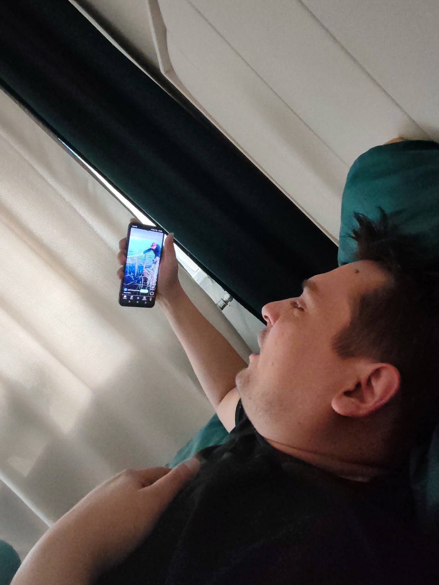 A man is depicted lying down, viewed from a slightly elevated perspective. He has short, dark hair and is wearing a dark t-shirt, resting his head on a dark teal pillow. He holds a black smartphone in his right hand, extending it upwards to look at the screen. The phone screen displays an image or video featuring a person, possibly wearing a red hat, in what appears to be an outdoor setting, potentially near water or on a wooden structure. Generic app icons are visible at the bottom of the phone screen, but specific text is illegible. The man's mouth is slightly open, indicating engagement with the content.
In the background, light-colored vertical blinds or curtains are visible, letting in natural light from what appears to be a window. A darker fabric element, likely another curtain or part of a window frame, is present to the right. The setting appears to be indoors, possibly a bedroom or living room, during daylight hours. The provided location context for this image is Podolsk, Russia.
