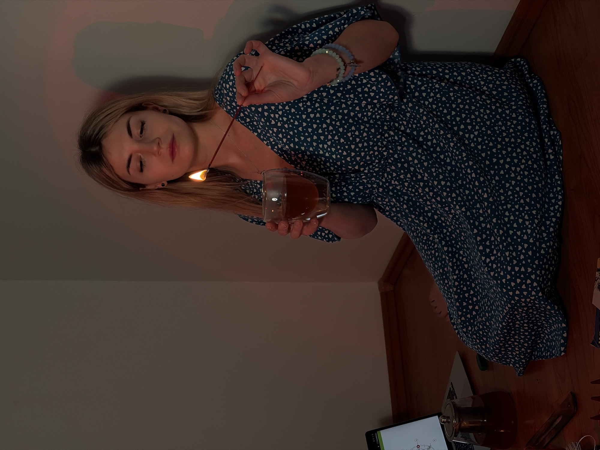 A young woman with blond hair, wearing a blue dress with white hearts, is positioned in the corner of a room. She is holding a clear glass mug filled with a dark liquid, potentially tea, and an incense stick that is lit at the end. She is also wearing bracelets. The setting is indoors, likely in a residential space, with a laptop and teapot on a wooden floor. The location is unspecified, but the query mentions the city of Budva, Montenegro, as context.