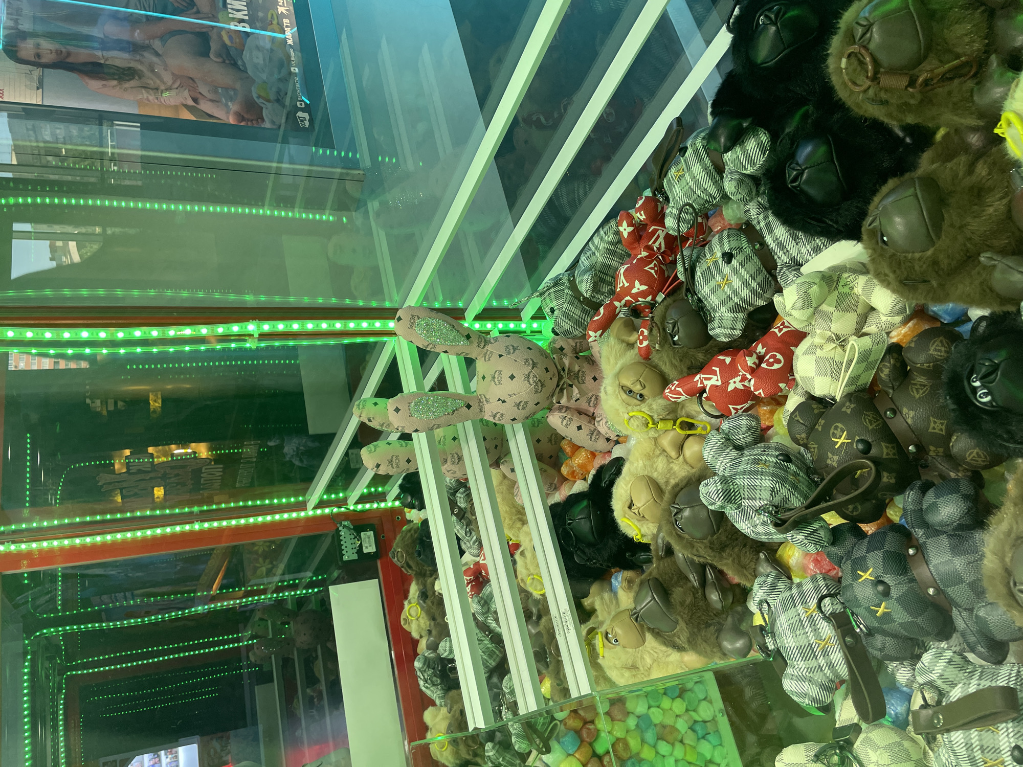 The image shows the interior of a claw machine filled with plush toys. The toys are predominantly ape-like figures and some other animal-themed plushies. Many of the ape figures have faux fur and some are wearing sunglasses. Several of the plush toys are patterned with what appears to be a luxury brand's monogram, such as Louis Vuitton. In the center of the machine, a large pink plush rabbit with green glitter accents on its ears is prominently displayed. The claw machine is illuminated by bright green LED lights, creating a vibrant and somewhat futuristic ambiance. In the background, through the glass of the machine, a movie poster is partially visible. The scene is set inside what appears to be an arcade or entertainment venue. There are no people clearly visible interacting with the machine in the frame. The location is identified as Vaganovo, Russia.