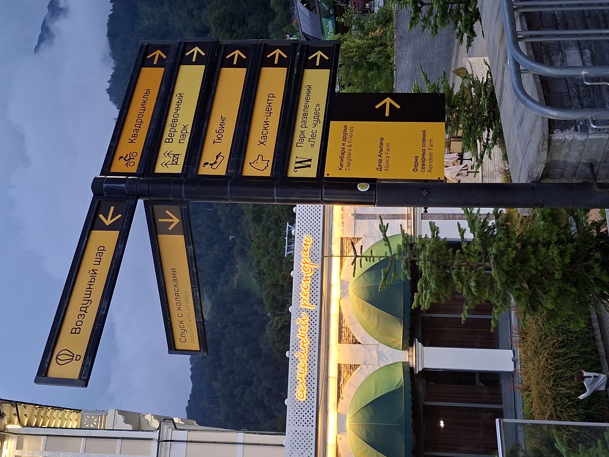 The image shows a directional signpost with multiple yellow signs pointing in various directions, indicating different attractions. The signpost is located in Estosadok, Russia, as evidenced by the Russian text on the signs. One sign says "Семейный ресторан" which translates to "Family restaurant" in English, and it is illuminated with neon lights, suggesting it is a restaurant.
The signs on the post offer directions to various activities and locations:
- "Воздушный шар" (Hot air balloon)
- "Спуск с колясками" (Descent with strollers)
- "Квадроциклы" (ATVs)
- "Веревочный парк" (Rope park)
- "Тюбинг" (Tubing)
- "Хаски-центр" (Husky center)
- "Парк развлечений «Лес чудес»" (Amusement park "Wonderland")
- "Капибара и друзья" (Capybara and friends)
- "Дача Альпача" (Alpaca Farm)
- "Ферма северных оленей" (Reindeer Farm)
The environment appears to be a mountainous resort area, with a forested mountain slope visible in the background under a cloudy sky. The foreground shows a building with arched awnings, a stone wall, and evergreen trees. A person is partially visible in the lower left, near some bushes. The overall scene suggests a recreational area or resort offering a variety of outdoor and family-friendly activities.