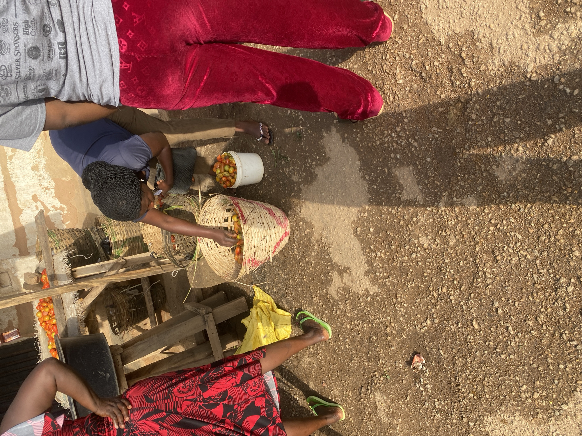 This image depicts a vibrant street scene in Jos, Nigeria, likely in the late afternoon given the long shadows cast on the dusty, gravelly ground. The subjects are three women, their clothing suggesting a casual, everyday setting. One woman, wearing a grey t-shirt and dark pants, is partially visible at the top left. Her clothing has some printed text, including "SILVER SPONSORS HIGH Craft." Another woman, in the center, wears a dark blue t-shirt and has her hair covered in a black net. She is actively engaged, reaching into a woven basket filled with red tomatoes. The third woman, on the right, is wearing bright red, patterned leggings and green flip-flops. She appears to be sitting or squatting.
The primary activity is the preparation or display of produce, specifically tomatoes, which are overflowing from a woven basket and also contained in a white bucket. Wooden structures, possibly market stalls or makeshift shelving, are visible in the background, adding to the market-like atmosphere. A bright yellow plastic bag lies on the ground near the woman in red leggings, contrasting with the earthy tones of the surroundings. The overall mood appears to be one of daily commerce and activity, with the focus on the fresh produce. The sun is casting strong shadows, indicating it's likely a sunny day.