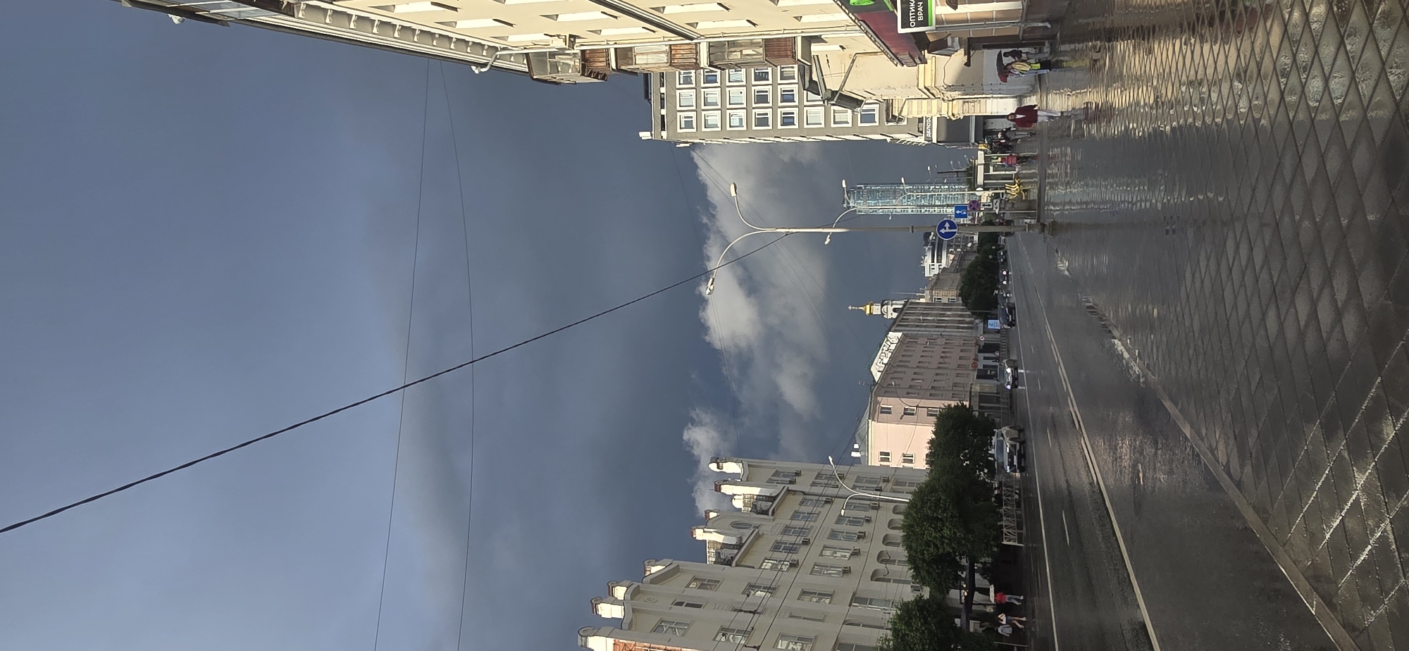 This image captures a wide city street in Yekaterinburg, Russia, under an overcast sky marked by dark, heavy storm clouds. The asphalt road and adjacent tiled sidewalks are wet and reflective, indicating recent precipitation. The street extends into the distance, accommodating multiple lanes of traffic. Several vehicles, including cars and a white SUV, are present on the wet roadway.
On the left side of the street, multi-story buildings with light-colored facades and decorative architectural elements line the street. Green trees are planted along the sidewalk in front of these buildings. A few individuals are walking on the left sidewalk.
On the right side, a multi-story building with balconies and a storefront displaying Cyrillic text ("ОПТИКА ВРАЧ") is visible. Further down, a modern, grey building with a grid of square windows precedes the prominent, tall, slender, blue-tinted Vysotsky Business Center skyscraper in the background. Several pedestrians are present on the right sidewalk; one individual in a red jacket walks towards the foreground, and a group further away includes a person holding a red umbrella.
Overhead, a network of power lines or tram lines crisscrosses the sky. The cloud cover is predominantly dark and grey, with a slightly lighter patch of sky visible towards the upper left. Street lamps with double light fixtures are positioned along the street, and a blue circular traffic sign indicating a mandatory right turn is visible on a pole on the right side. In the mid-ground on the left, a pale pink building is visible, and behind it, a building topped with a golden-domed spire can be seen.