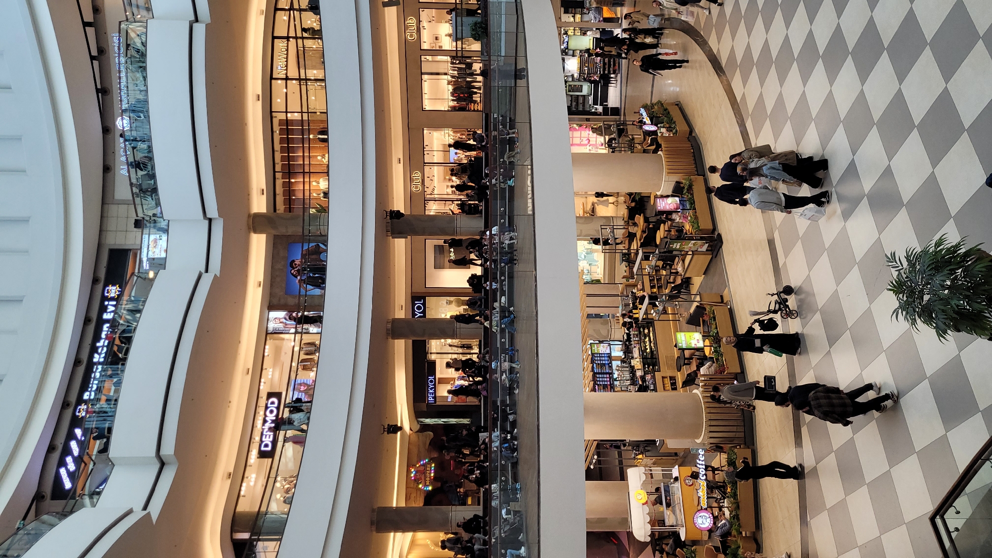 This is an indoor scene of a multi-level shopping mall. Several retail stores with glass fronts and signs (e.g., "DER MOD", "IPEKYOL", "Net Work", "Bursa Kahan Evi") line the different levels, and there is a food court area. People are seen strolling on the tiled floor and seated at the food court tables. The shopping mall has large, white, curved architectural features. The image appears to be taken from an elevated point of view looking down on the scene. The shopping mall is in Başakşehir, Turkey.