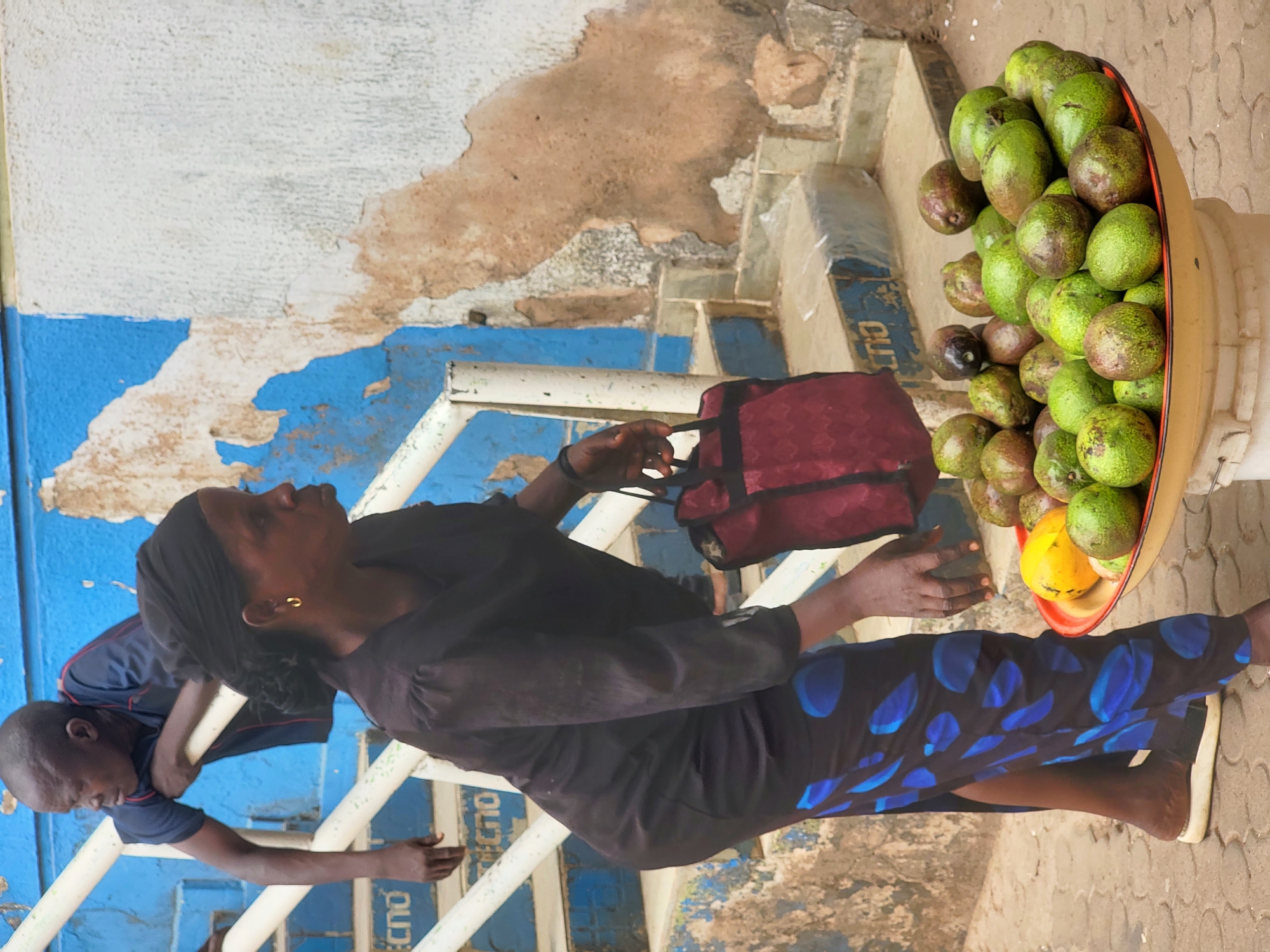 The photograph, taken in Jos, Nigeria, captures a vibrant street scene centered around a fruit vendor. The image is oriented vertically (originally taken in portrait mode and then rotated), showing a woman in the foreground and a man/boy in the background, set against a backdrop of a weathered wall and a paved ground.
In the right foreground, a woman with dark skin stands beside a large, yellow basin overflowing with fresh green avocados. A bright yellow mango is also visible among the avocados. The woman is dressed in a dark, long-sleeved top and a long skirt or dress featuring a distinctive blue and black leaf-like pattern. She wears a black headscarf and a gold earring in her right ear. Her left hand holds a dark red bag, while her right hand is near the fruit display. She looks upwards and to the left, her expression not clearly discernible. She appears to be barefoot or wearing light-colored flat footwear.
Behind her, on the left side of the frame, a man or older boy with dark skin is partially visible. He wears a dark blue short-sleeved shirt, possibly with red trim, and is leaning against or holding onto a white metal railing. His head is cast downwards, looking out of frame.
The background features a wall with considerable peeling paint, revealing layers of white, bright blue, and a warm brown or beige underneath. The ground appears to be a light-colored, possibly paved or concrete surface. Visible text on one of the white railings reads "TECNO," a brand of mobile phones popular in the region. Another partial word, possibly "CUTO," is seen above it. The scene is illuminated by natural daylight, suggesting it is daytime. The overall impression is a candid glimpse into daily life and commerce in a Nigerian urban setting.