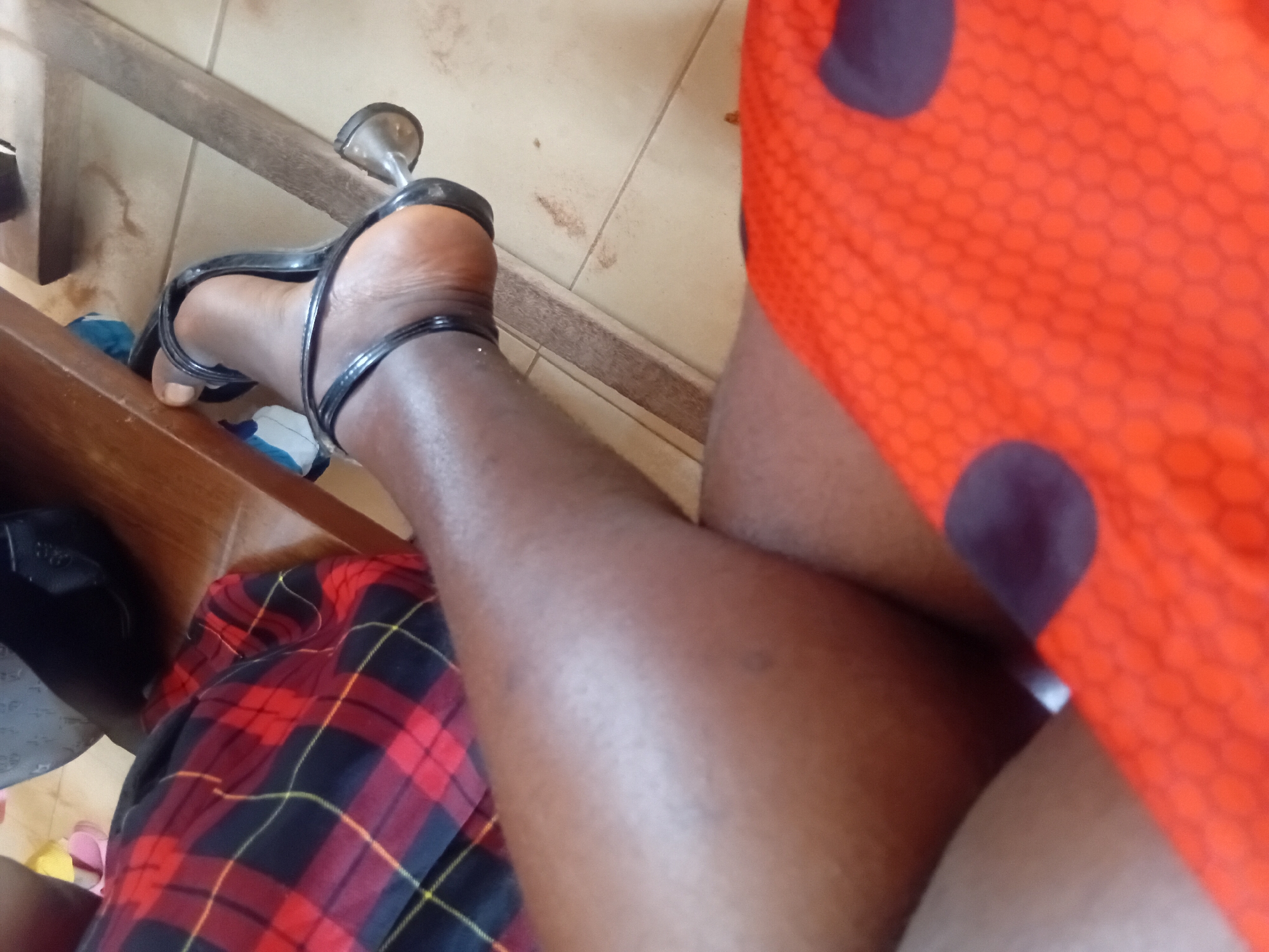 In Ejule, Nigeria, a person is seated, with their legs crossed, wearing a red and black plaid skirt and black high-heeled sandals. The person is also wearing an orange garment with a hexagonal pattern and dark purple circles, which partially covers their lap. A wooden table is visible, along with a portion of a black shoe and some scattered items on the floor. The scene appears to be indoors, possibly in a home or an office, given the presence of furniture and flooring. The lighting suggests it is daytime.