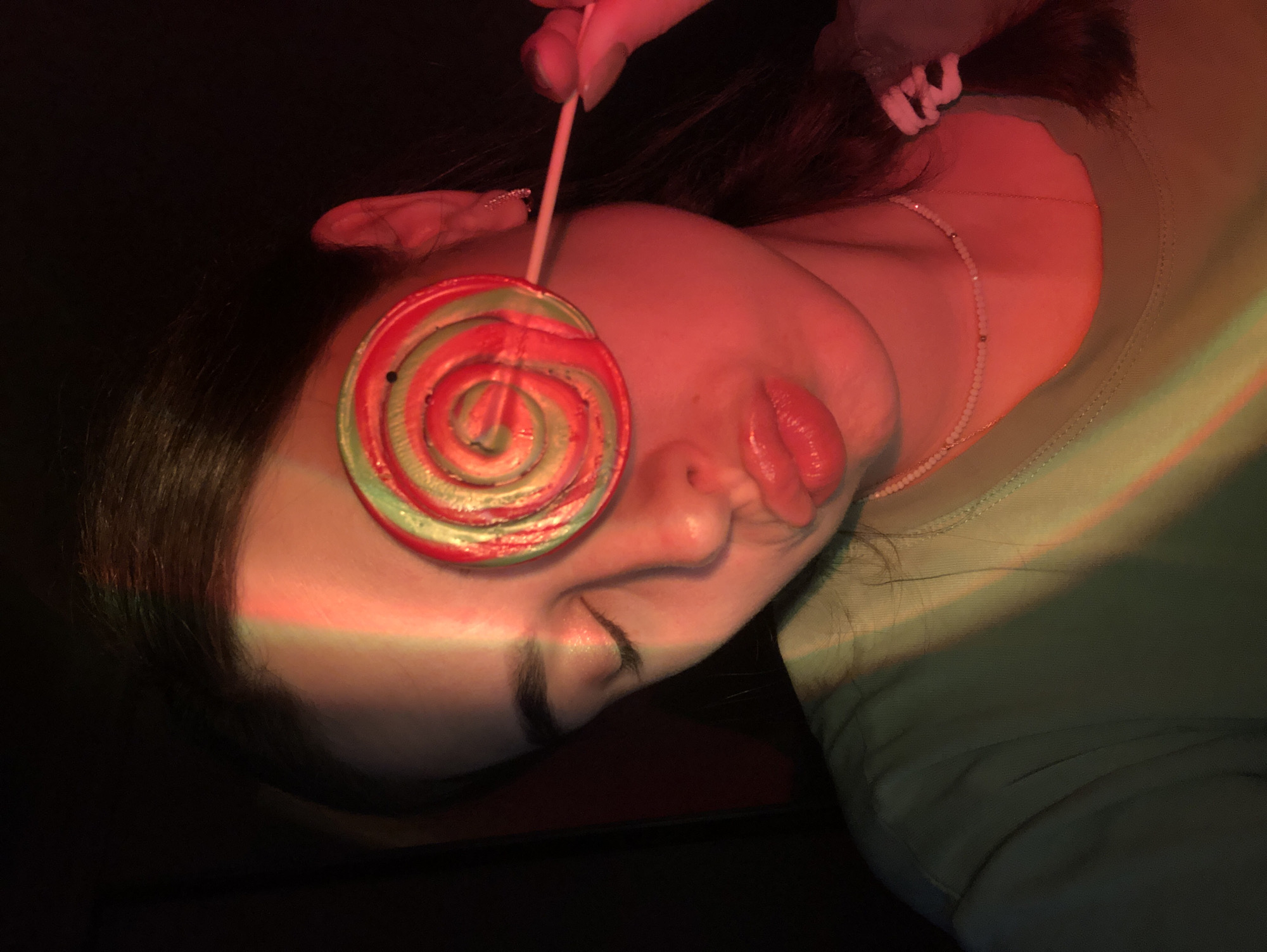 A young woman is shown in close-up, holding a red and green swirled lollipop in front of her right eye. Her left eye is closed, and her lips are pursed, as if she is about to kiss the lollipop. The scene is lit with red and green colored lighting, casting a colorful glow on her face and shoulder. She is wearing a light green t-shirt and a delicate beaded necklace. Her hair is dark and pulled back into a braid. The background is dark and out of focus. There is no information about the location in Noviy, Russia, or any other specific location.