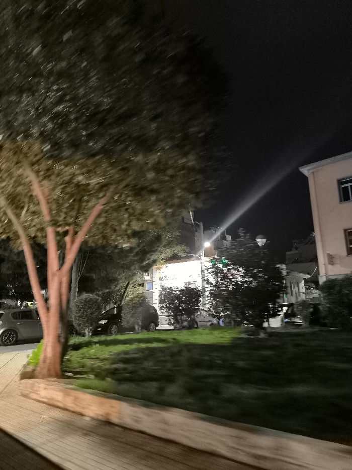This night-time photograph captures an urban scene in Drapetsona, Greece, illuminated by artificial lights. In the immediate foreground, a large tree with distinctive reddish-brown trunks and dark green foliage is prominently featured on the left, appearing blurred, which suggests motion or a shallow depth of field. A wooden or paved bench-like structure is visible along the bottom edge. To the right of the tree, a dark green grassy area fills the mid-ground.
Further back, several cars are parked or in motion behind a line of dense green bushes and smaller trees. Buildings are visible in the background. A brightly lit white building stands out in the center-left, from which a powerful beam of light emanates upwards into the dark, clear night sky. A green light source, possibly part of a sign, is also visible near the top of this white building. On its facade, there is some dark, illegible text against a white background, likely a business sign. To the far right, a lighter-colored building with a visible window is partially seen.
The time of day is night, with the sky appearing dark and clear. No specific human activity is discernible. The overall blurriness of the foliage and foreground elements contributes to an atmospheric quality, suggesting the photo might have been taken from a moving vehicle or with a slow shutter speed.