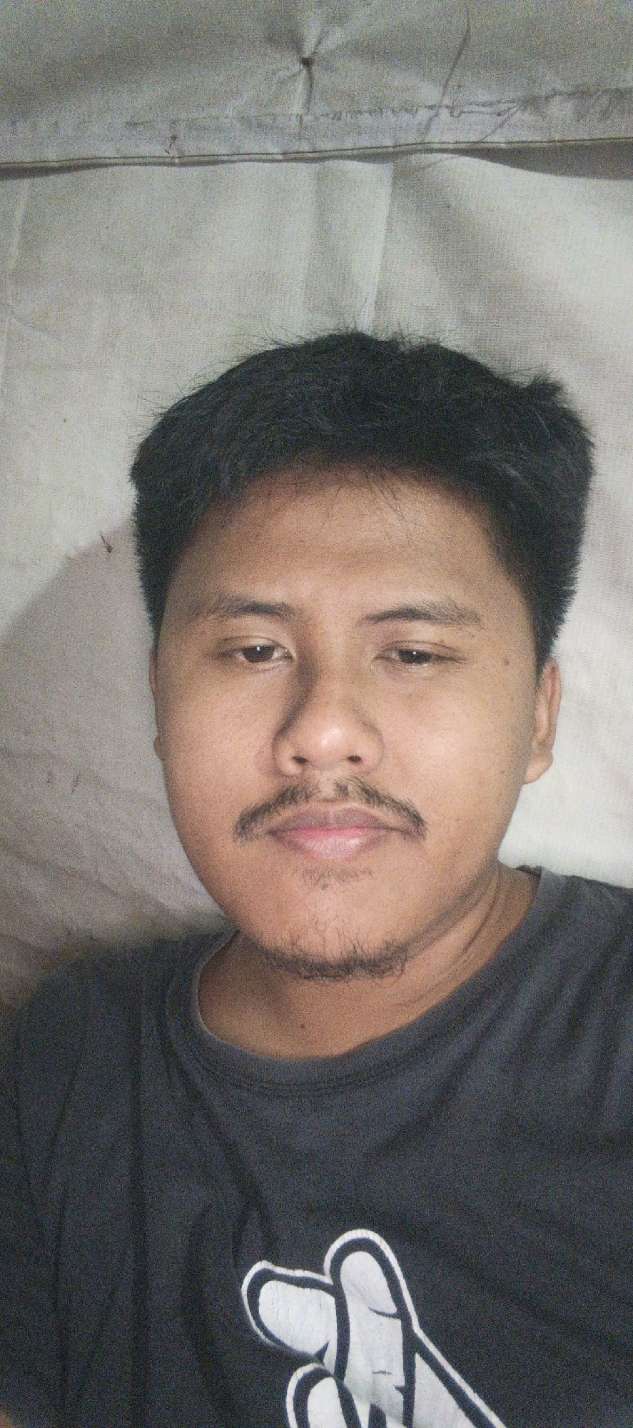 The image is a close-up, selfie-style portrait of a young East Asian male lying down on a light-colored, textured surface, likely a bed or mattress. The man has short, dark hair, prominent eyebrows, dark eyes, and a faint mustache and goatee. He is wearing a dark gray t-shirt with a white graphic design of two hands forming a shape, possibly a heart or peace sign. The background is a textured, off-white material, possibly a sheet or blanket, with a visible seam or fold running horizontally across the upper portion of the image. The lighting appears to be natural and diffused. The image is not clearly associated with the city of Kemuning, Indonesia, or any specific urban environment.