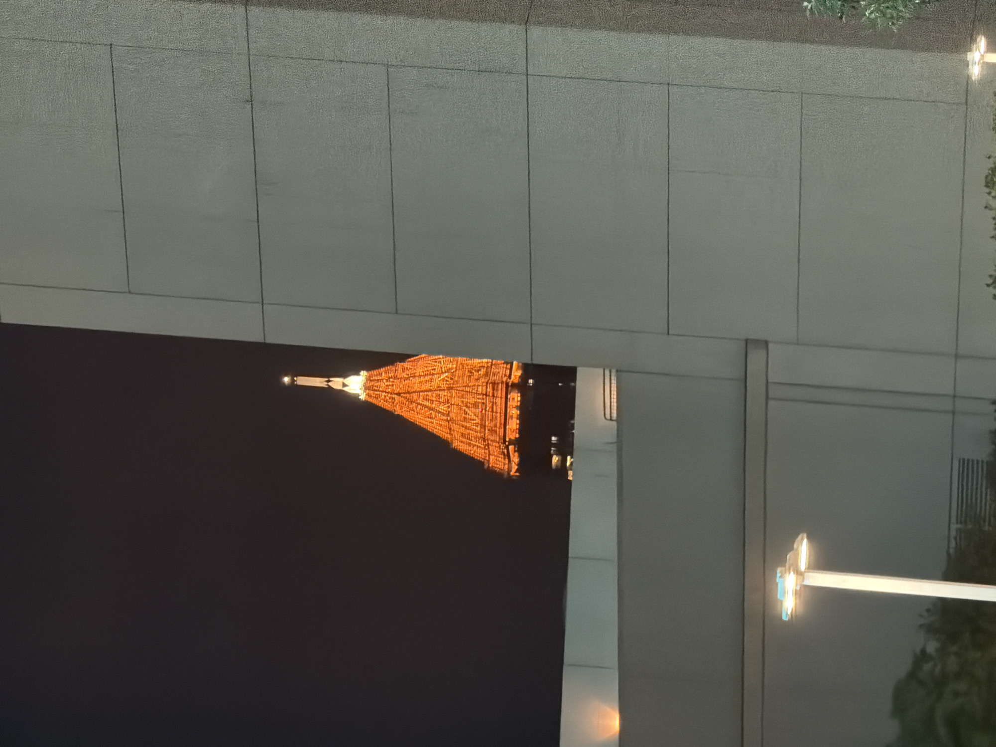 The image depicts the Tokyo Tower in Japan at night, viewed from an elevated position behind buildings. The tower is illuminated in orange light. A street lamp is also visible.